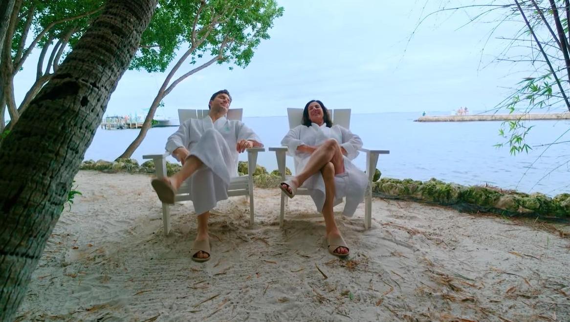 Two people relax in chairs on a sandy beach by the sea.