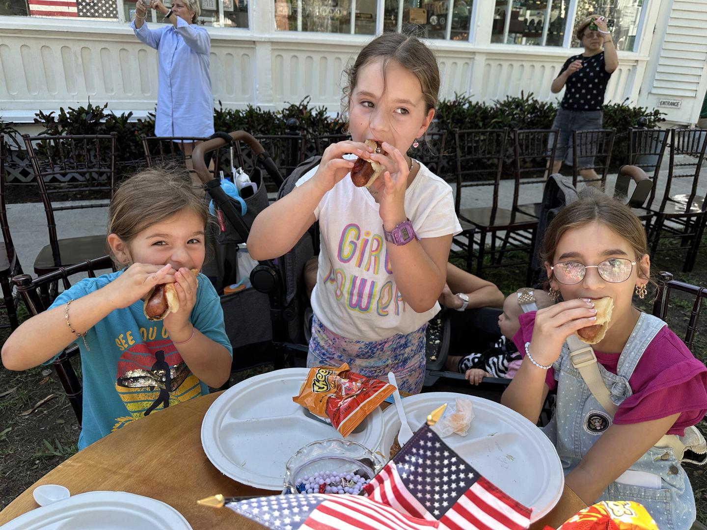 Three children eating hot dogs at a table with American flags.