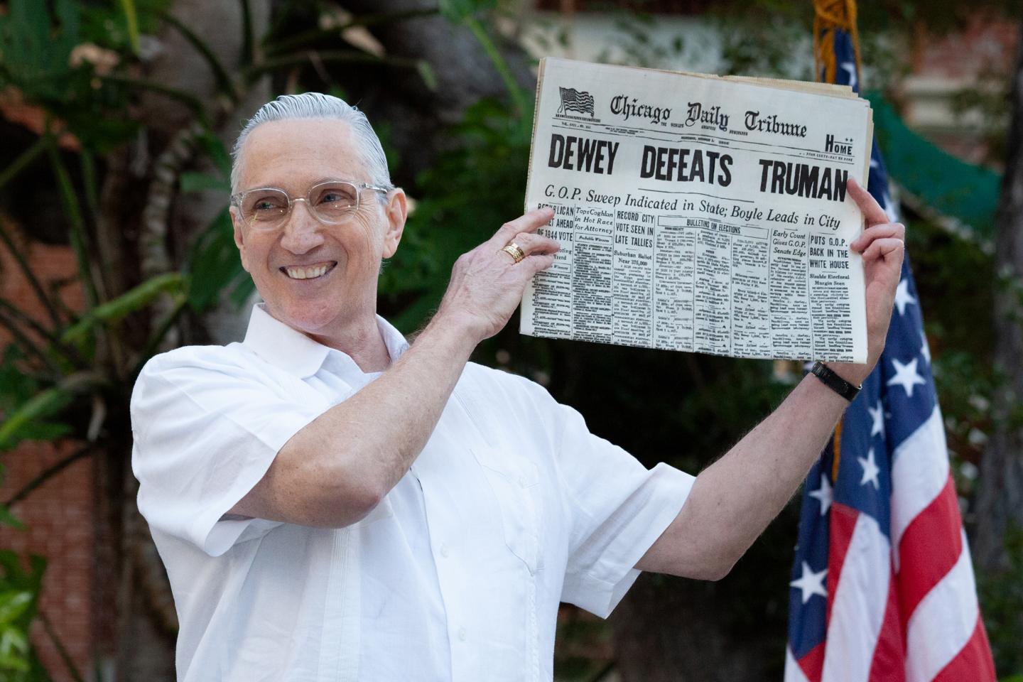 Smiling man holds up a historic newspaper; an American flag is nearby.