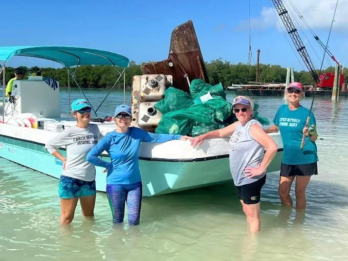 Four people stand beside a boat in shallow water, under a clear blue sky.