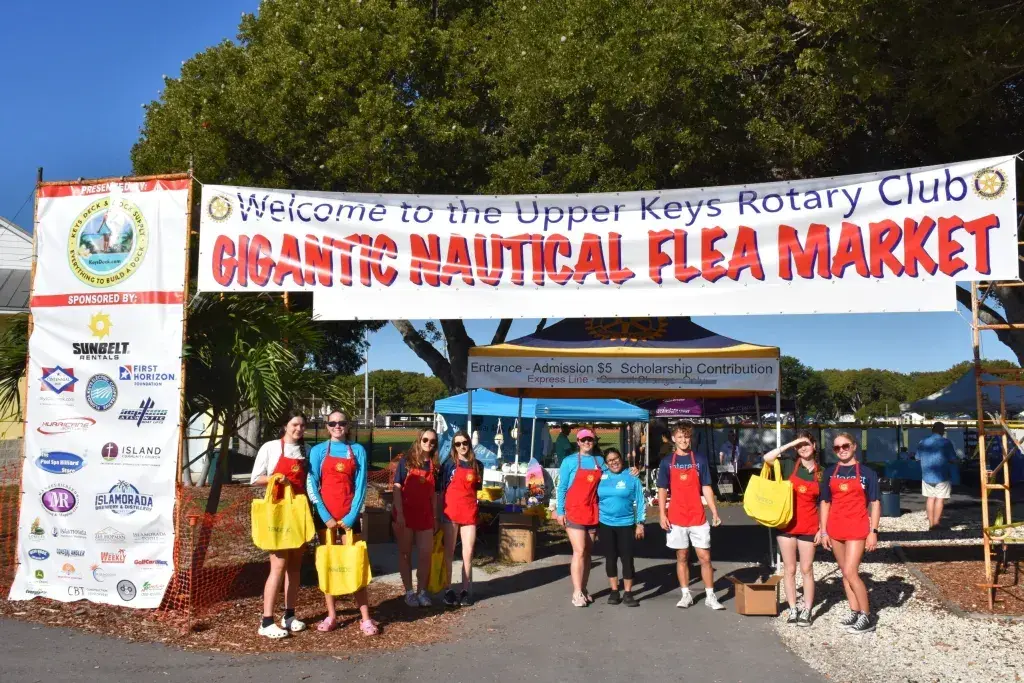 Volunteers stand under a banner for a nautical flea market on a sunny day.