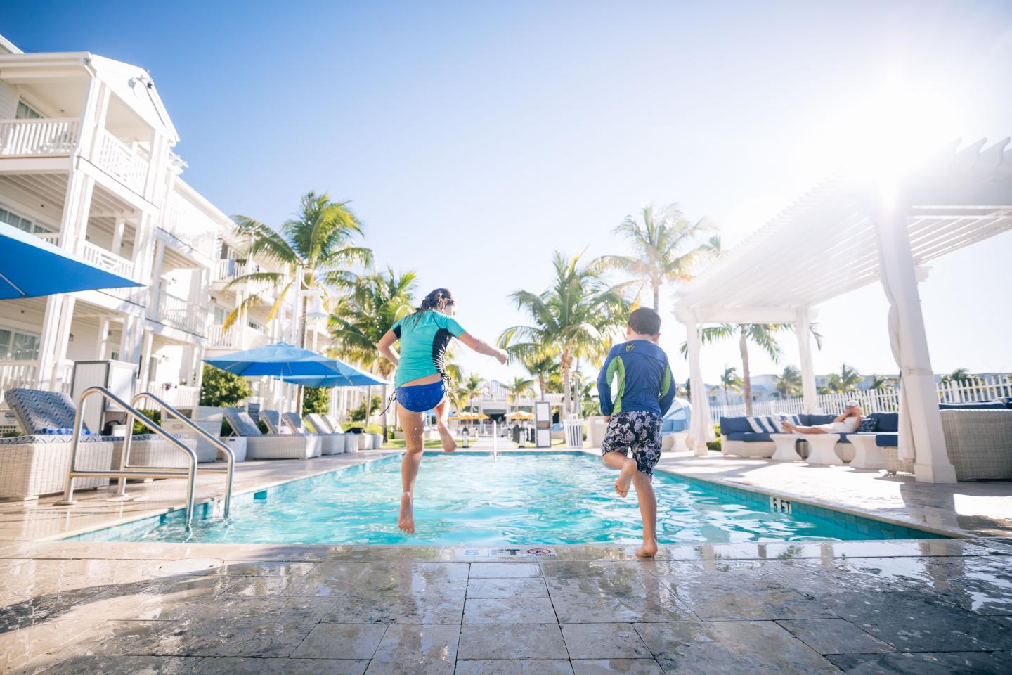 Children jumping into a sunlit pool at a resort with palm trees.