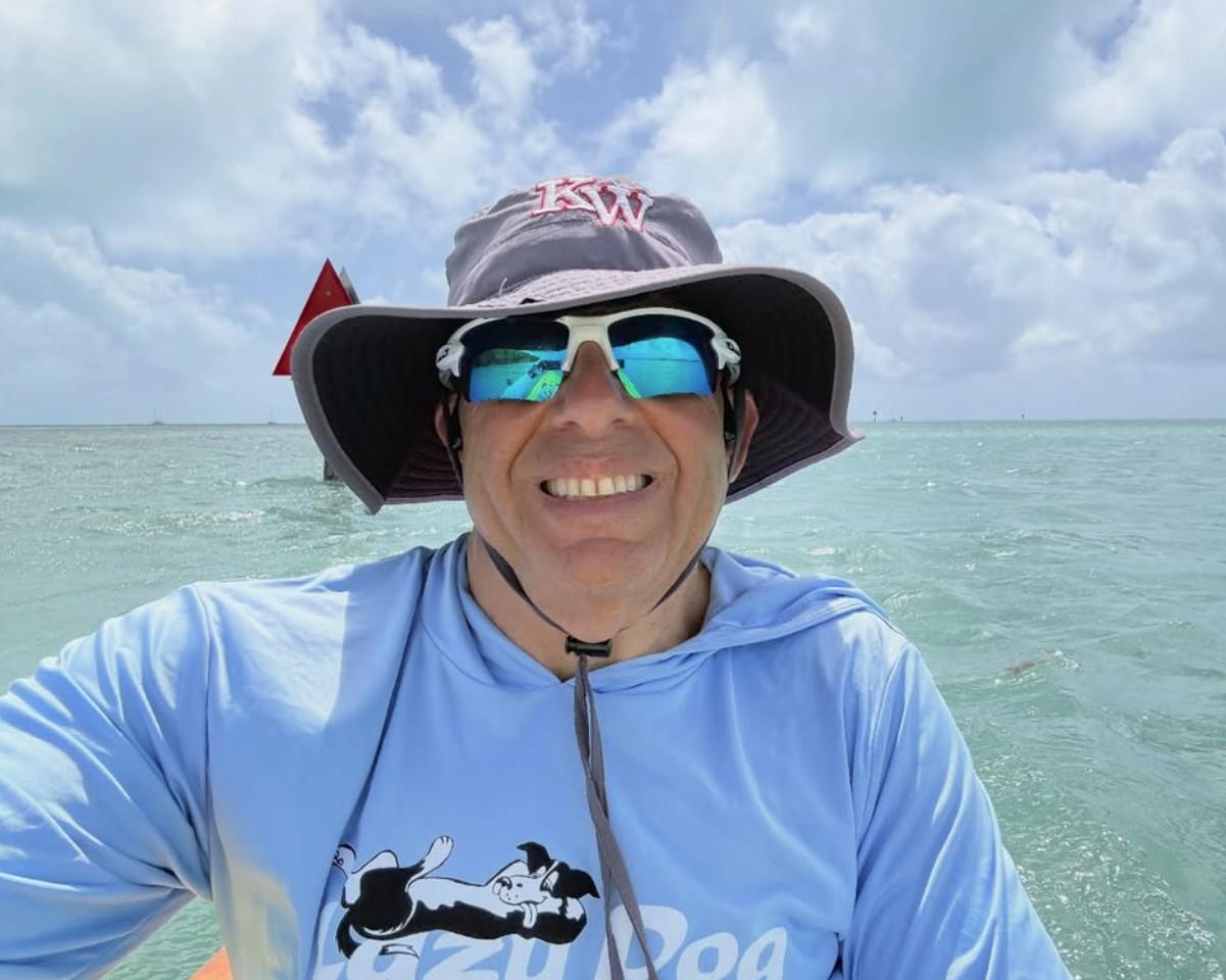 Man smiling on a boat in sunny weather, wearing sunglasses and a hat.