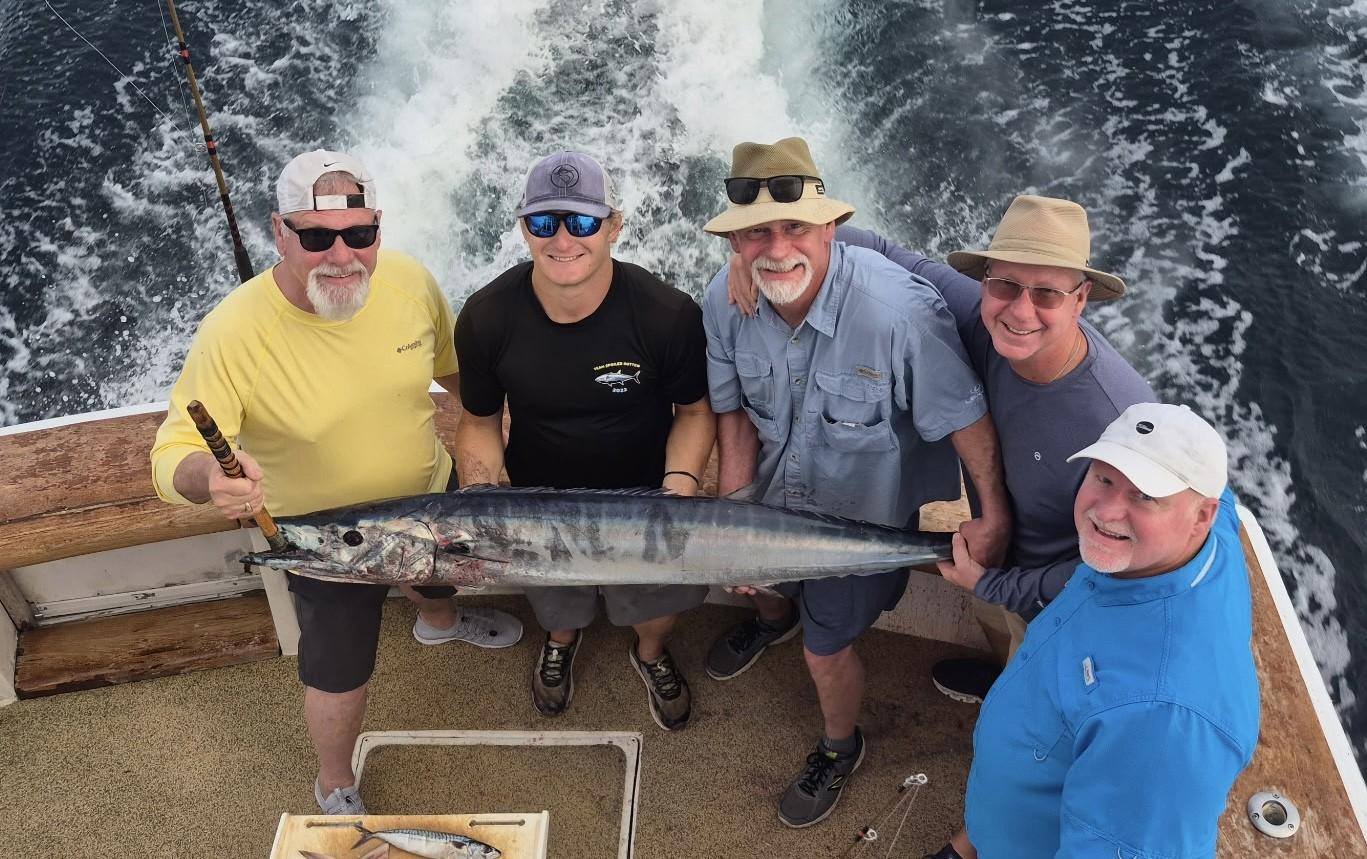 Five men on a boat holding a large fish, smiling, with ocean waves behind.