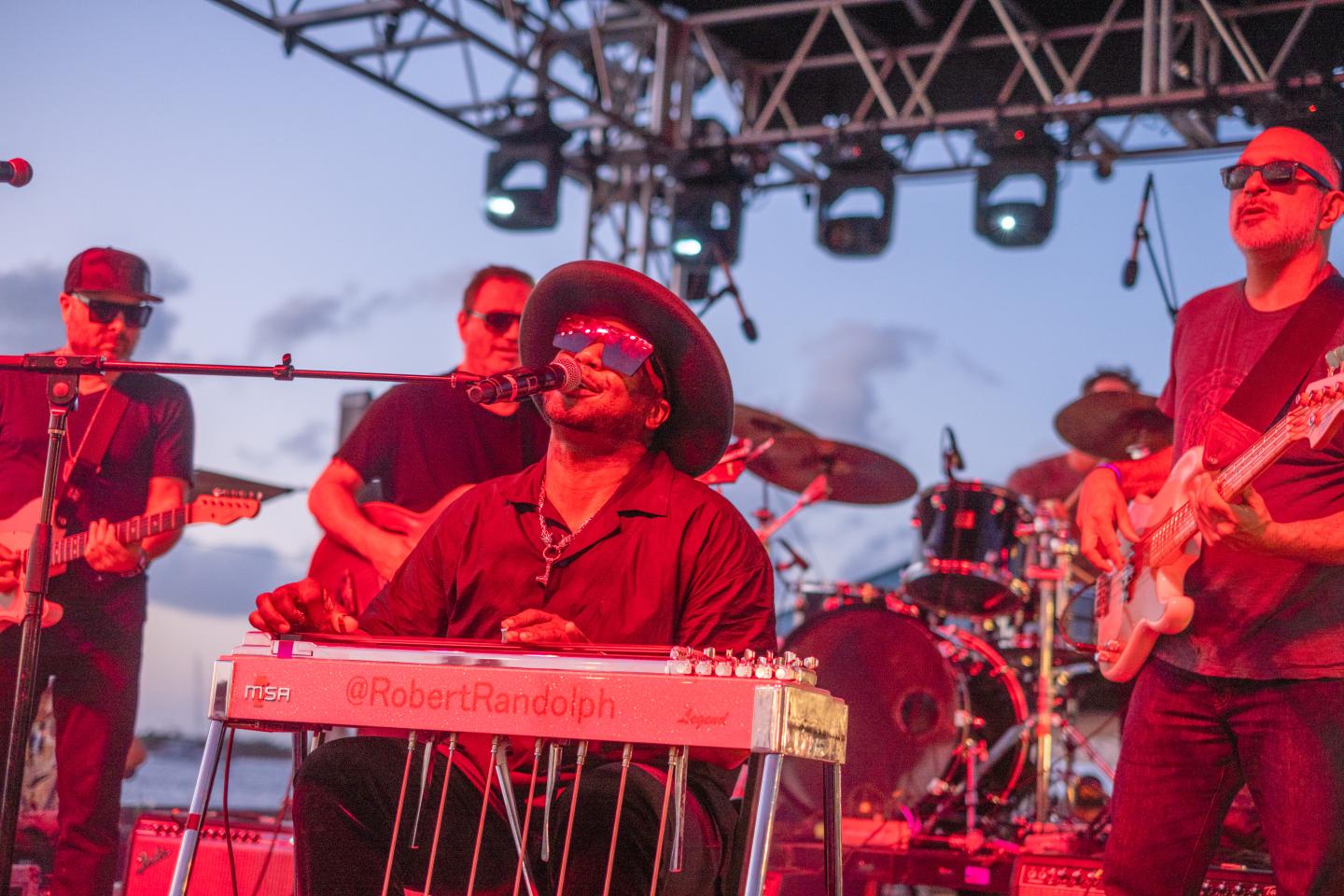Band playing on stage under red lighting, guitarist wearing a hat in the center.