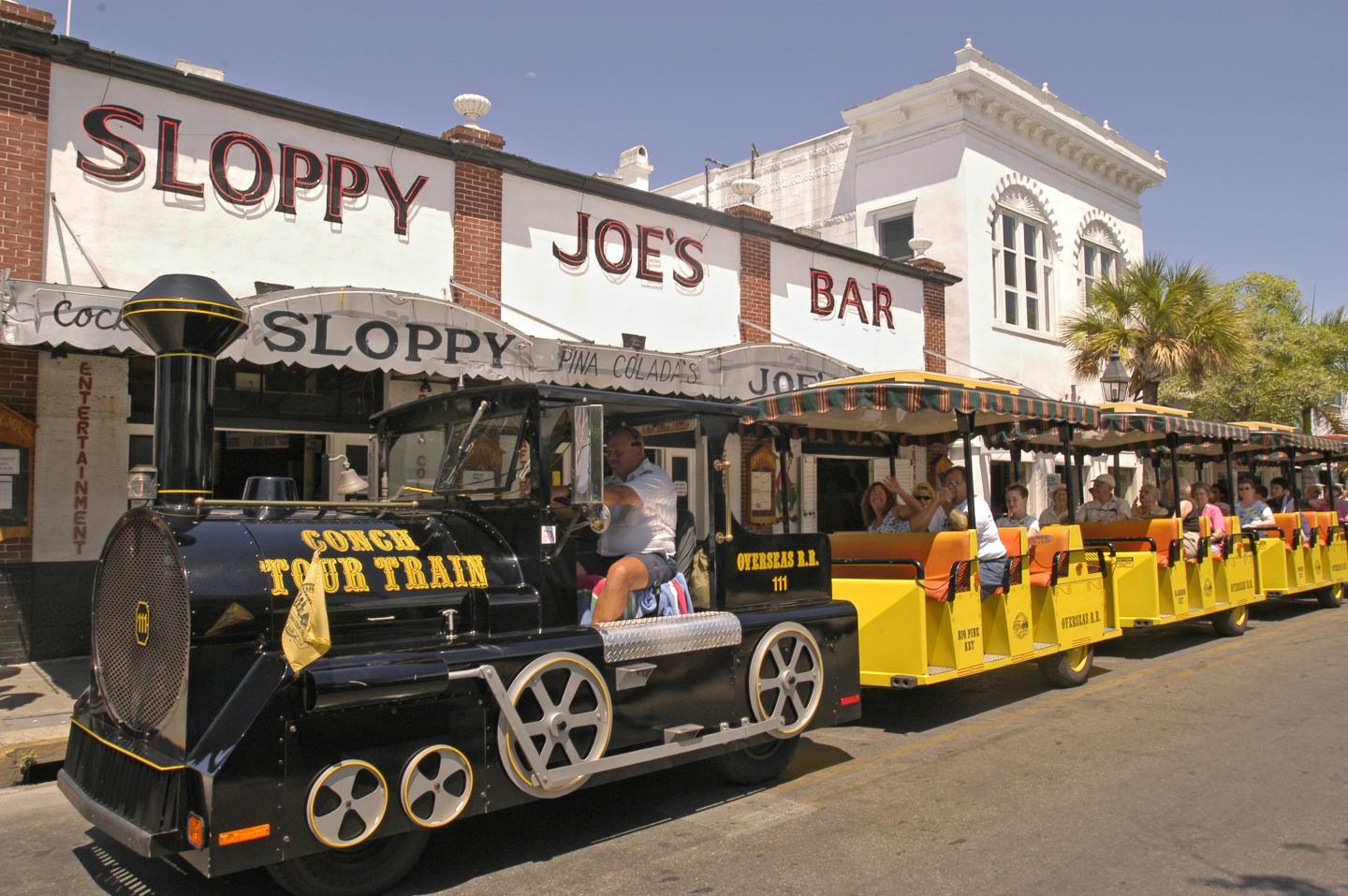 Tourist train passing a bar on a sunny street.