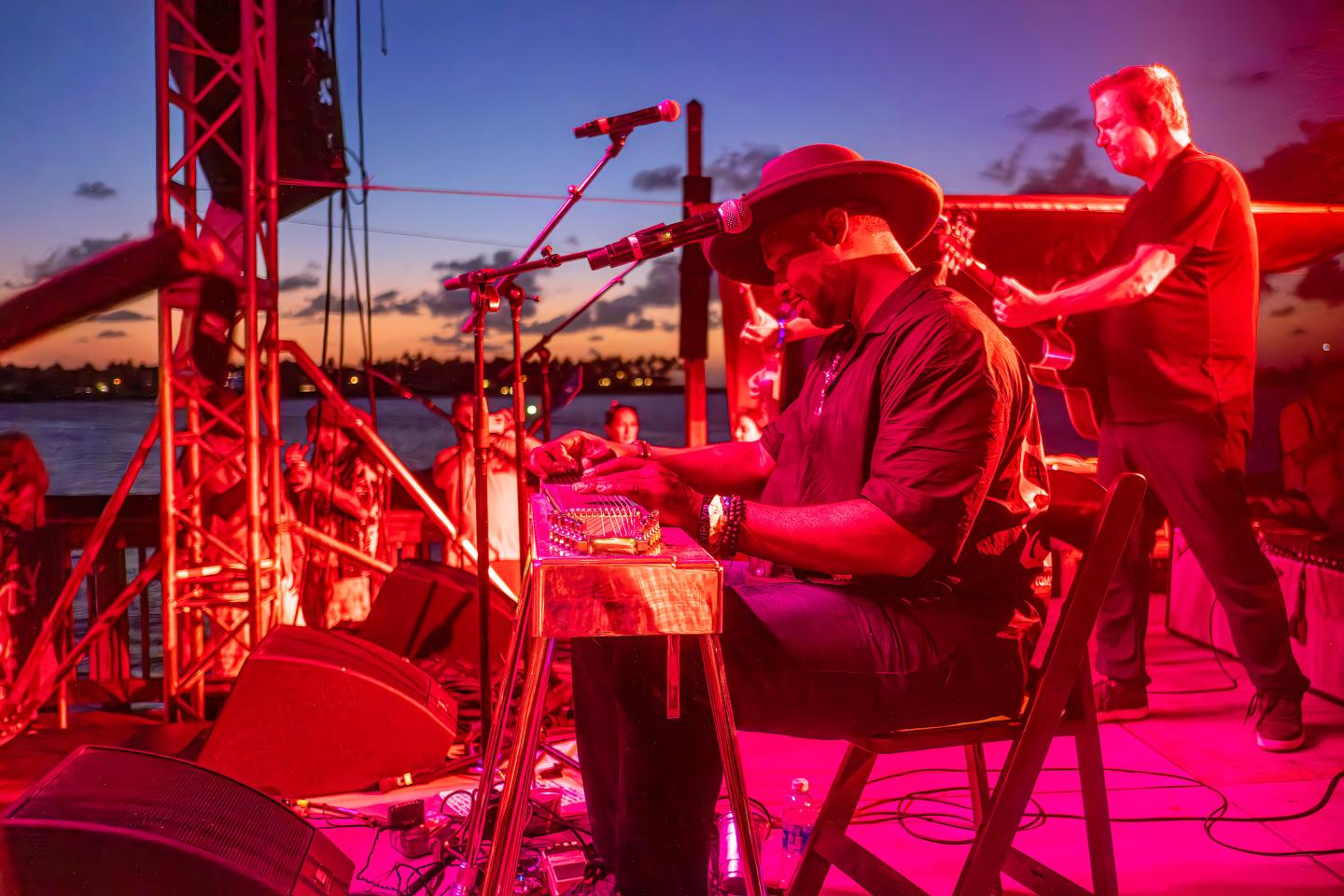 Musicians performing onstage at sunset, with vibrant red lighting.