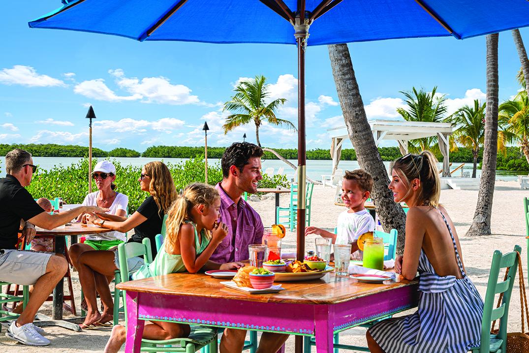 Family dining outdoors under blue umbrella, beach and palm trees in background.