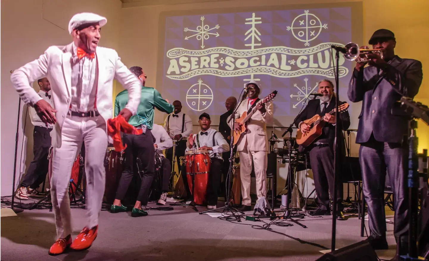 Dancer in white suit performs with a live band, vibrant stage lighting.