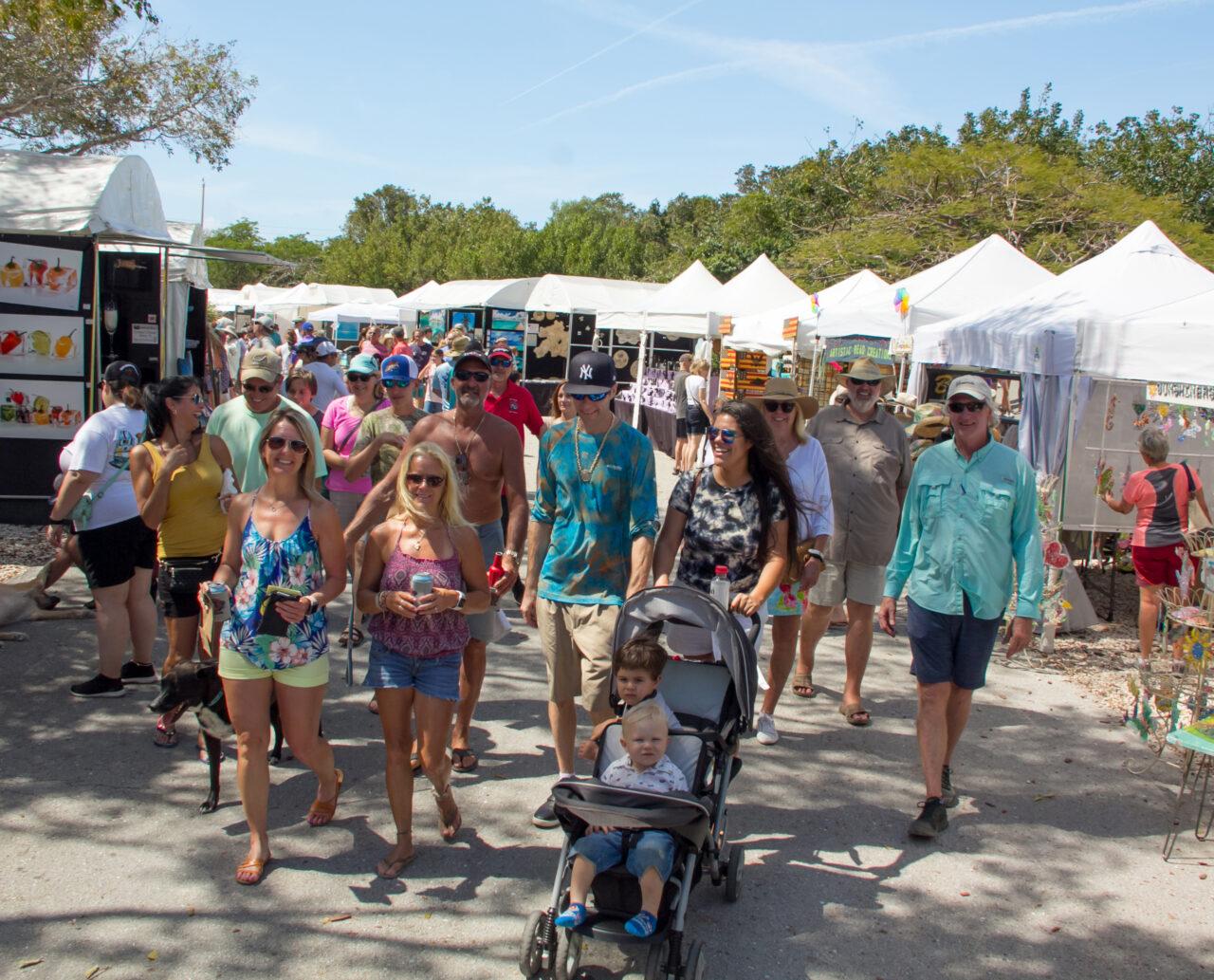 Crowd walking at an outdoor market with white tents and trees in the background.