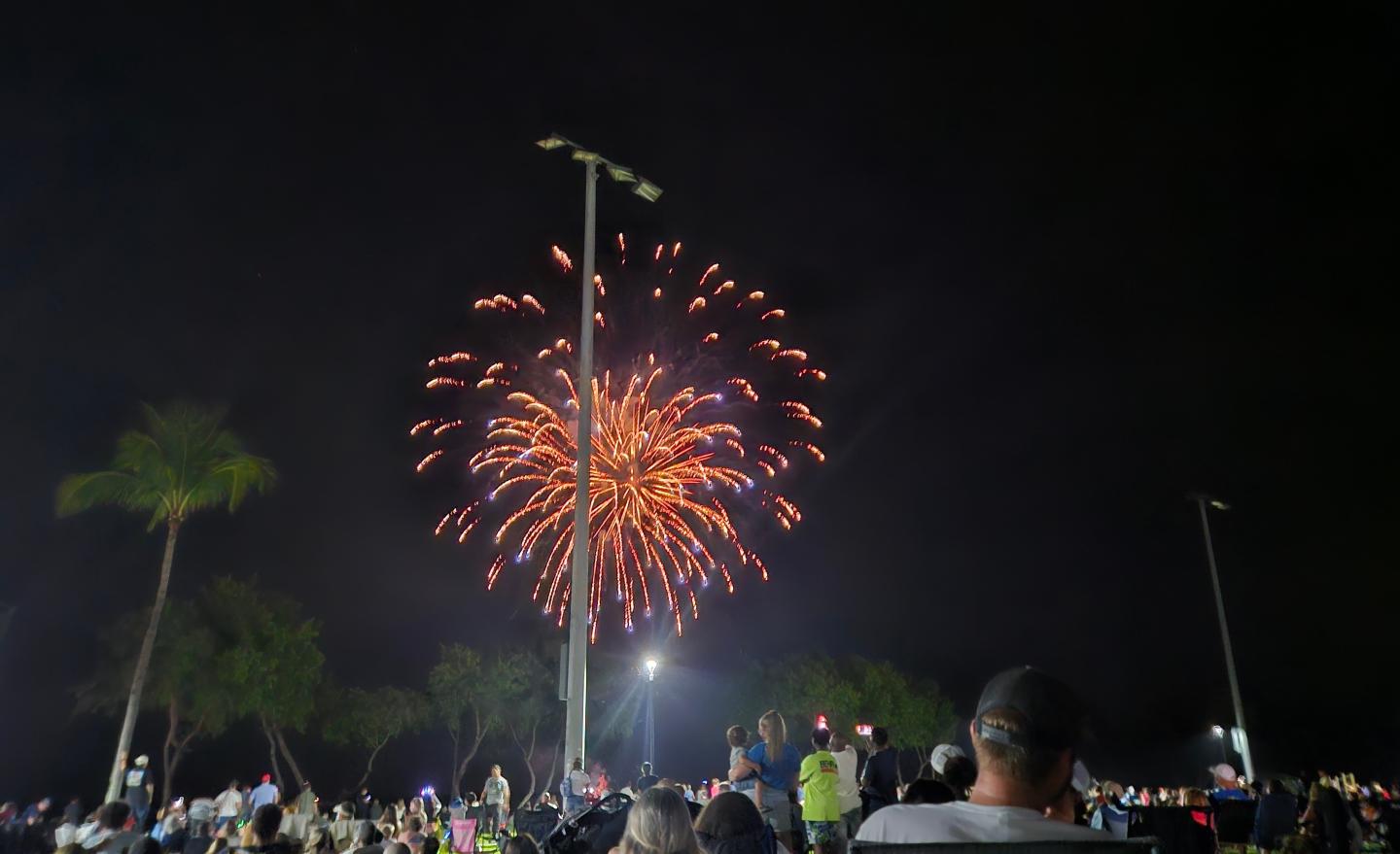 Fireworks light up a night sky over a crowd, palm trees visible.