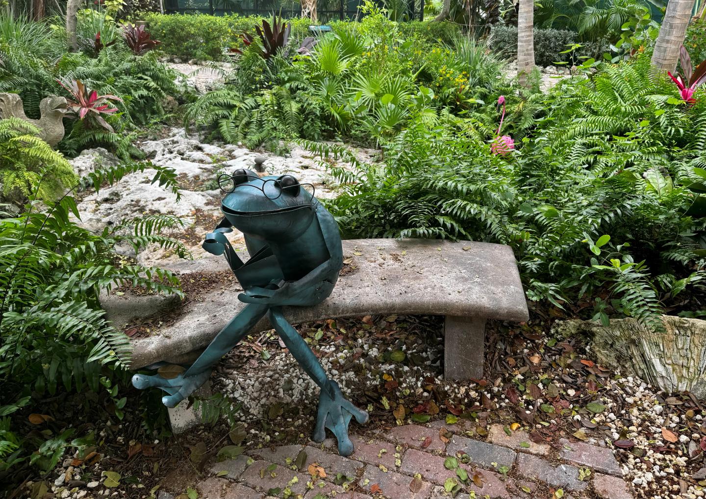 Statue of a relaxed frog on a garden bench surrounded by greenery.