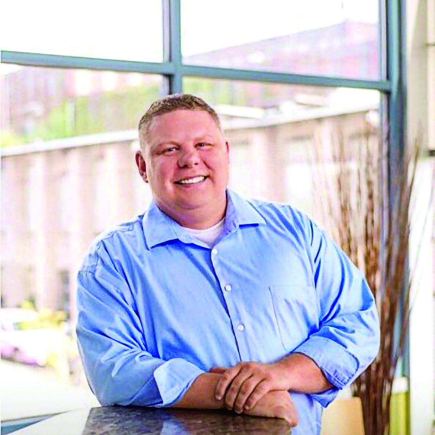 Smiling man in a blue shirt stands by a window.