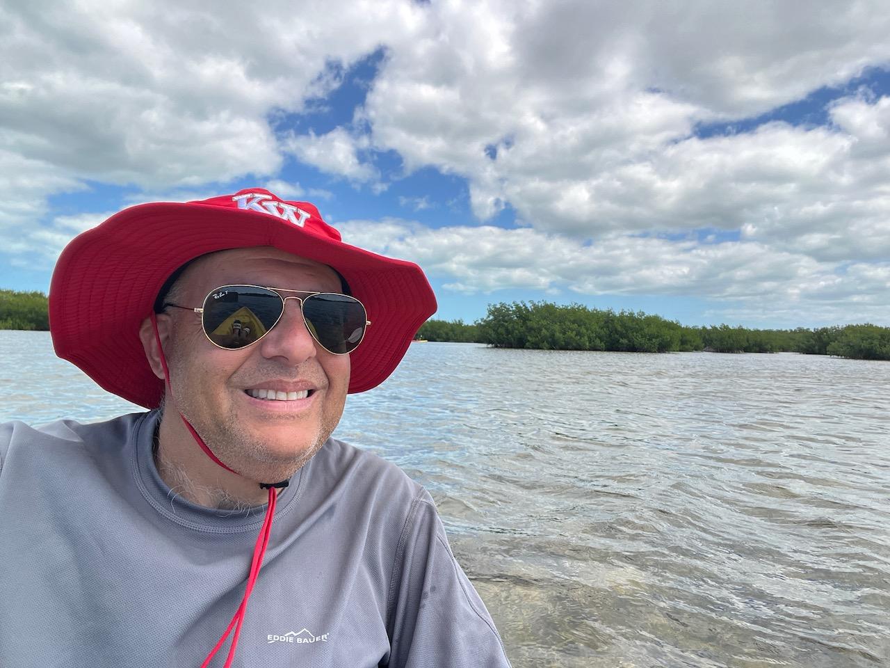 Man in a red hat and sunglasses smiles by a lake under a cloudy sky.