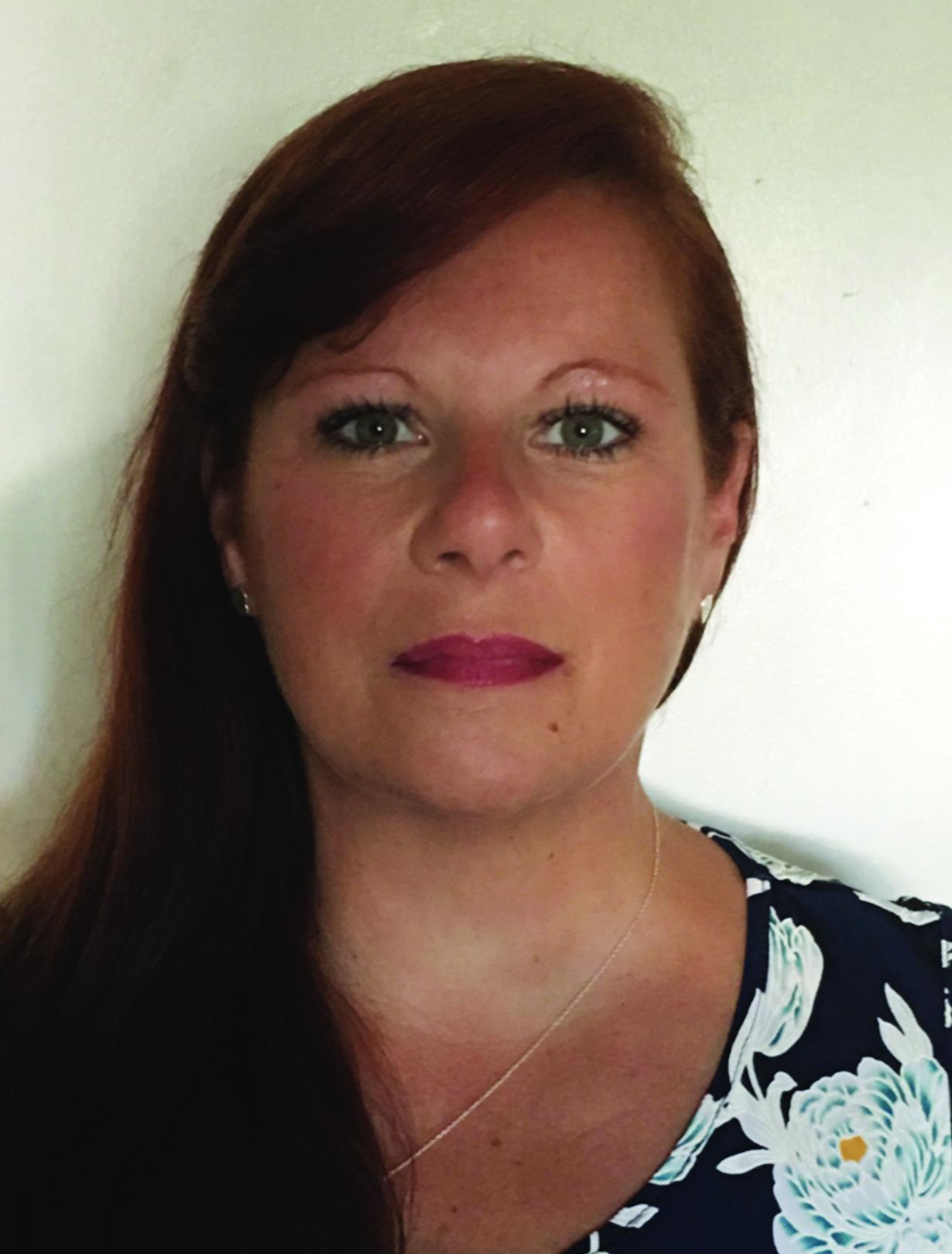 Woman with long red hair, wearing a floral dress, poses against a plain background.