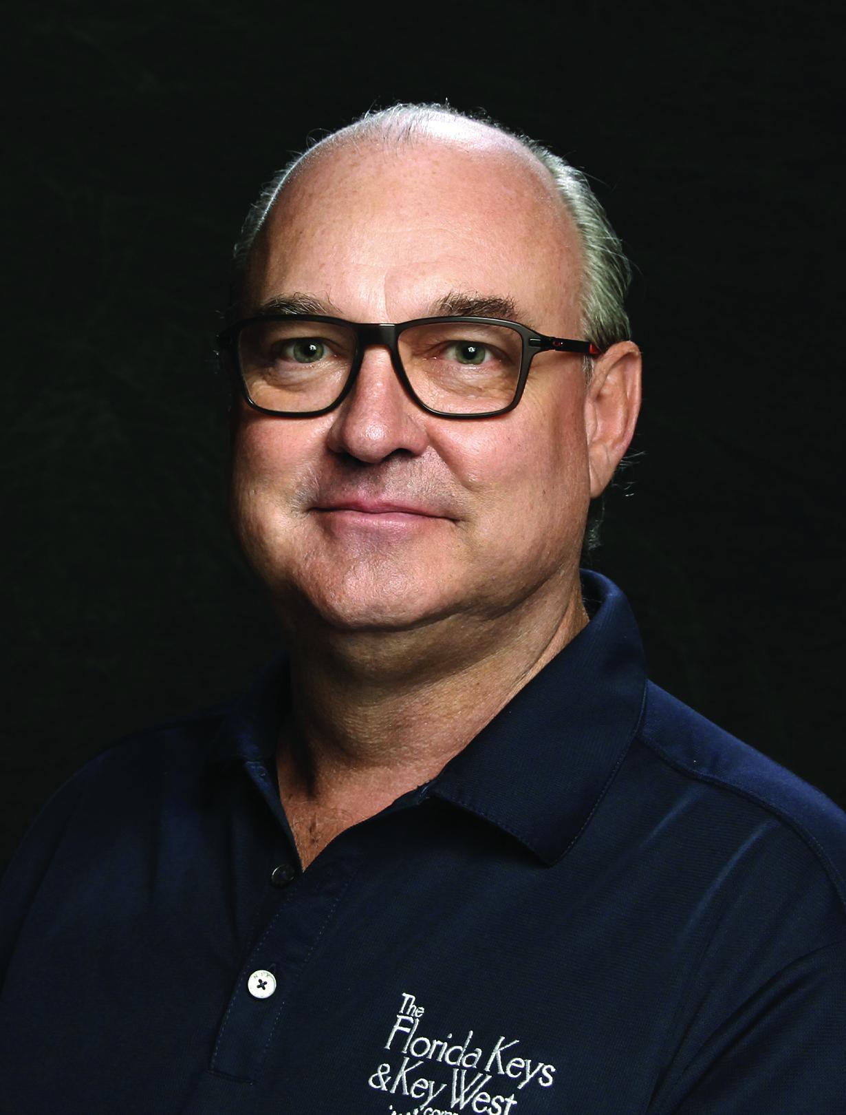 Older man with glasses and a navy shirt against a dark background.