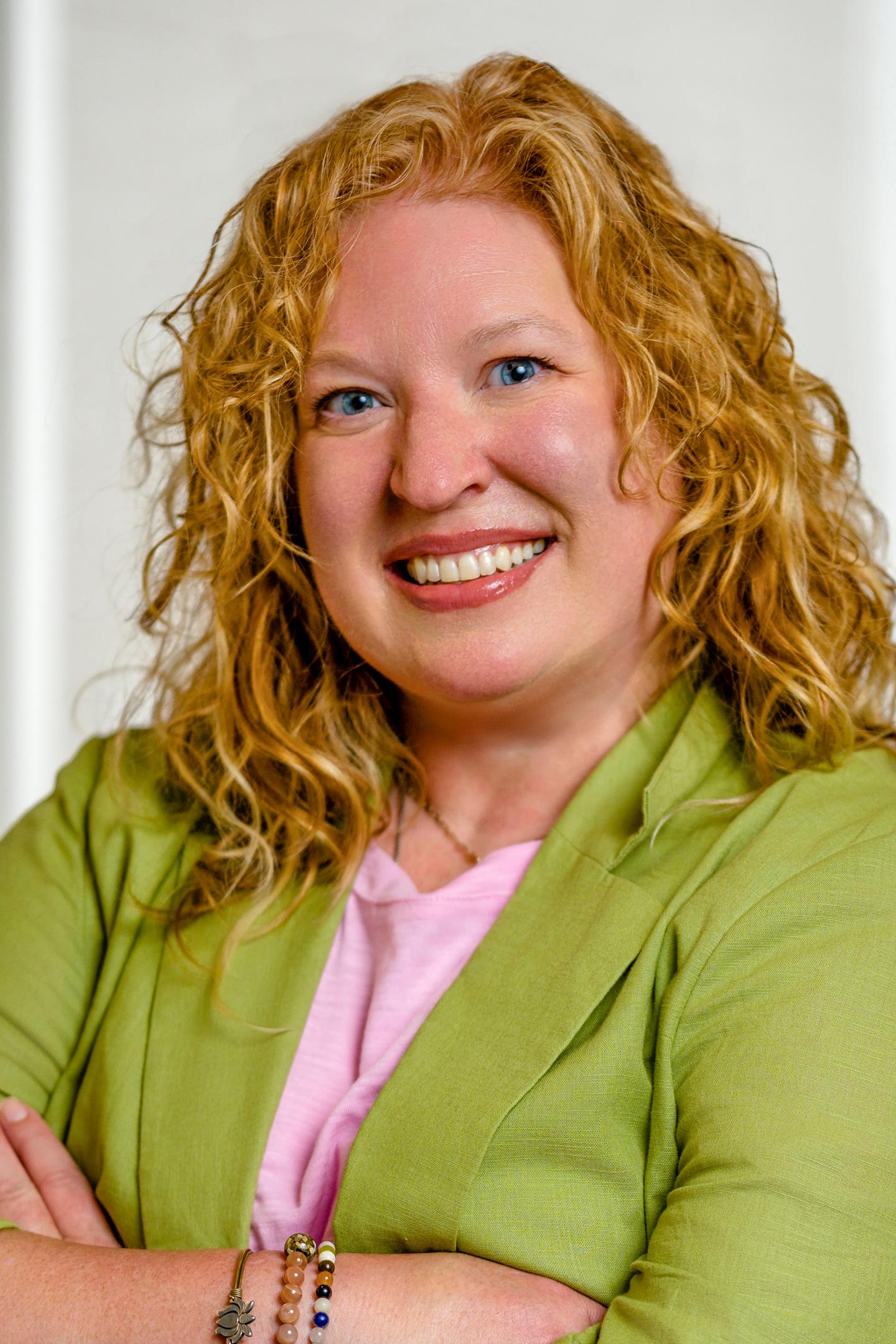 Smiling person with curly hair in green jacket and pink shirt.
