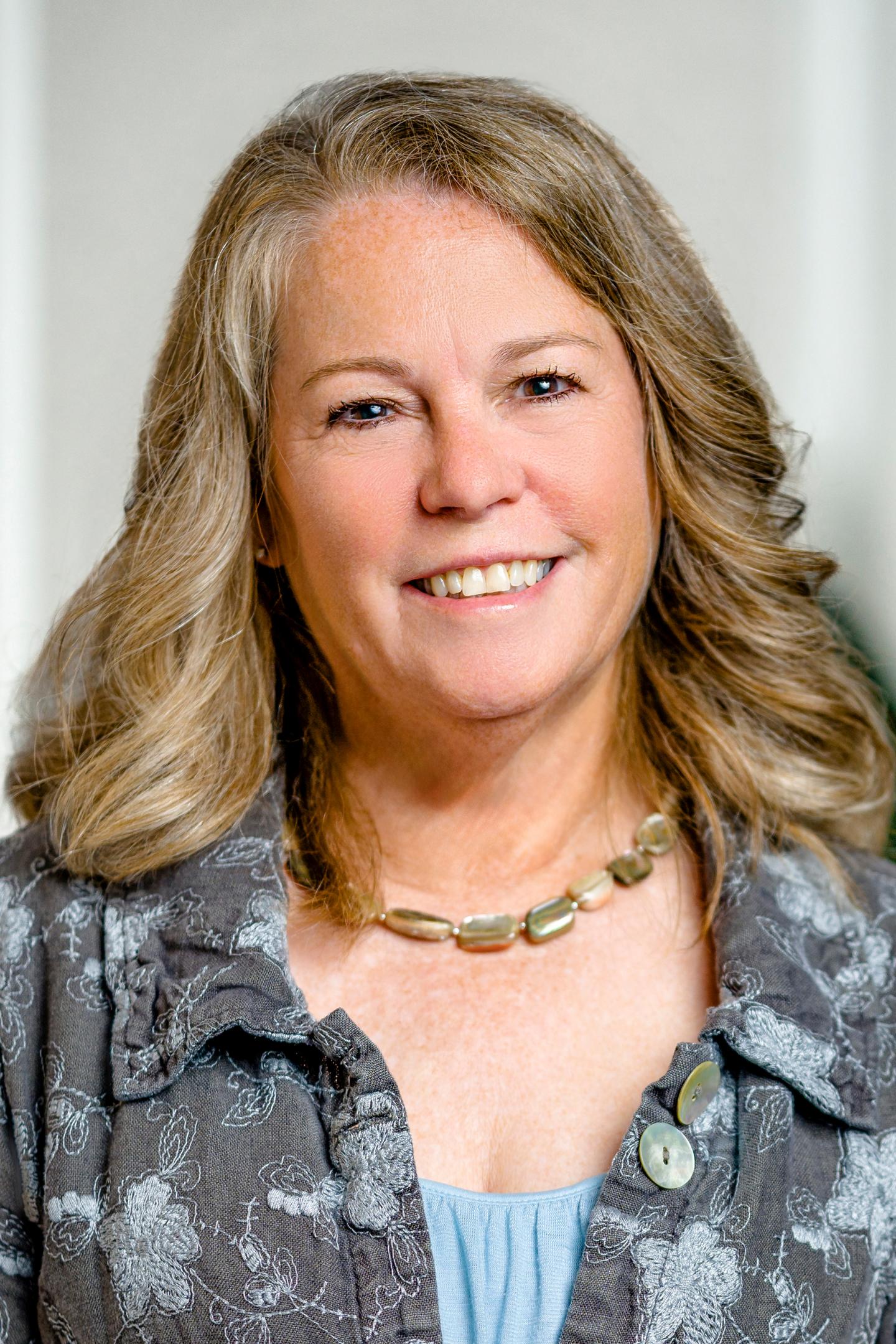 Smiling woman with long hair and a floral blouse, wearing a necklace.