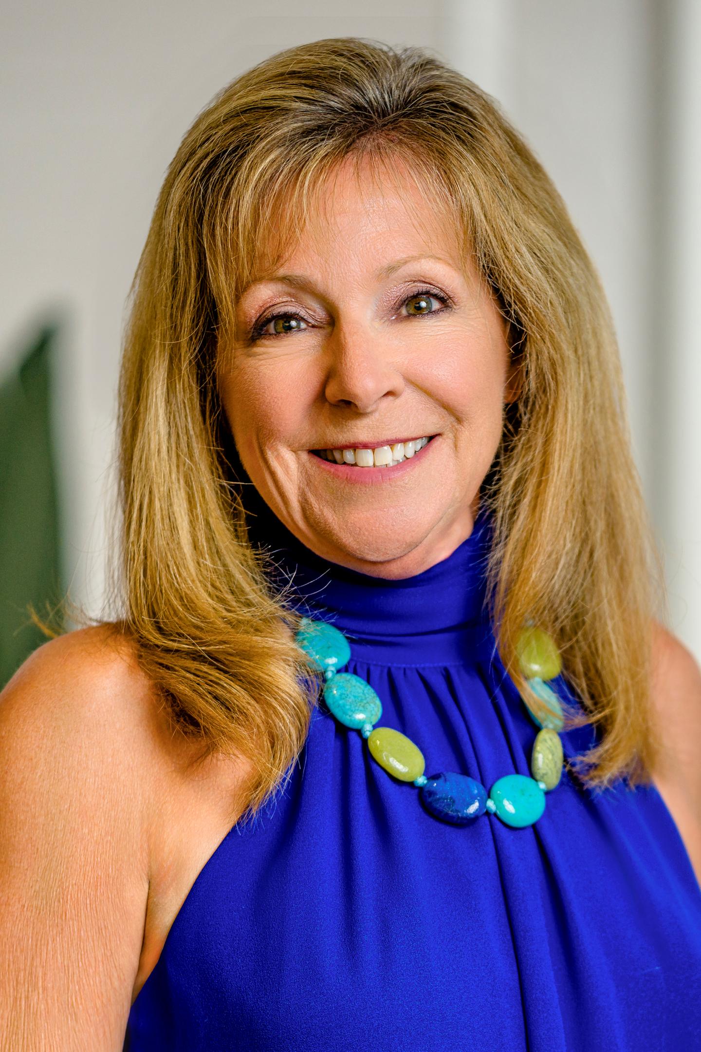 Smiling woman with long hair in blue top, wearing a colorful necklace.