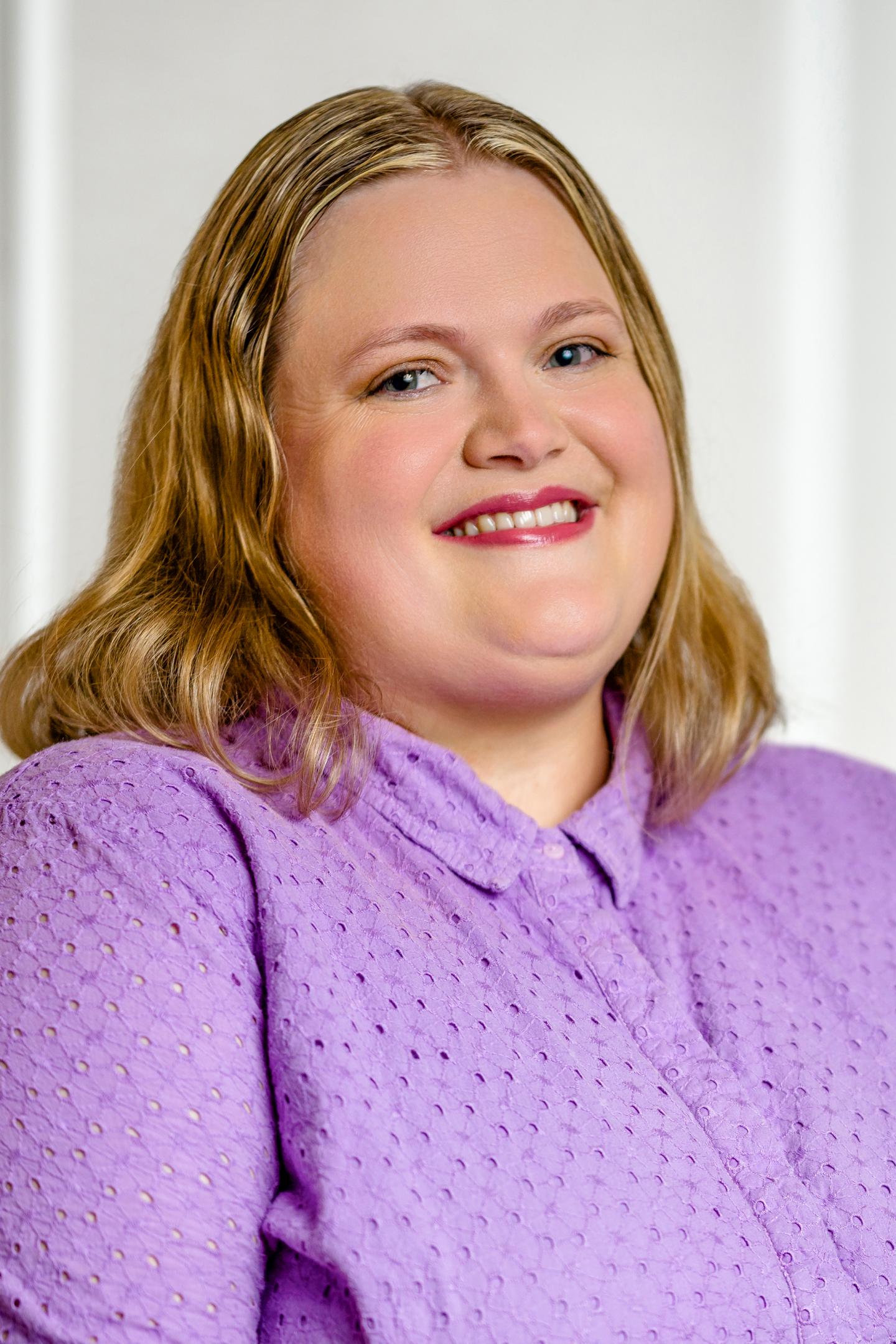 Smiling person in a purple blouse against a light background.