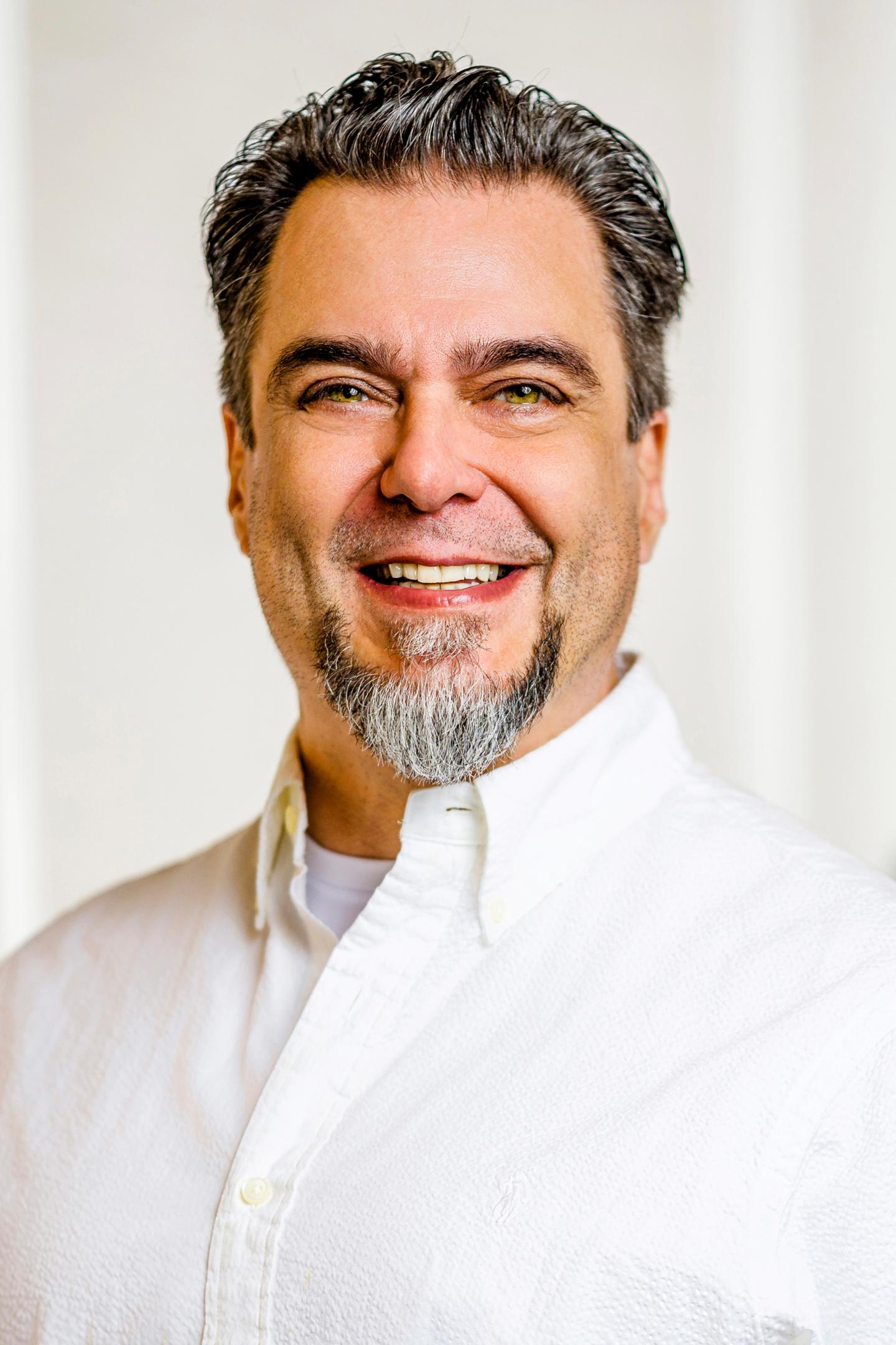Smiling man with a beard wearing a white shirt against a light background.