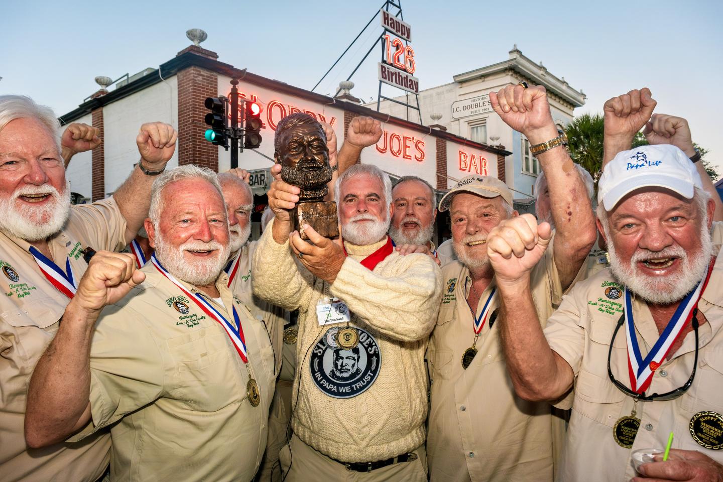 Group of bearded men celebrate outside a bar, holding up a bust trophy.