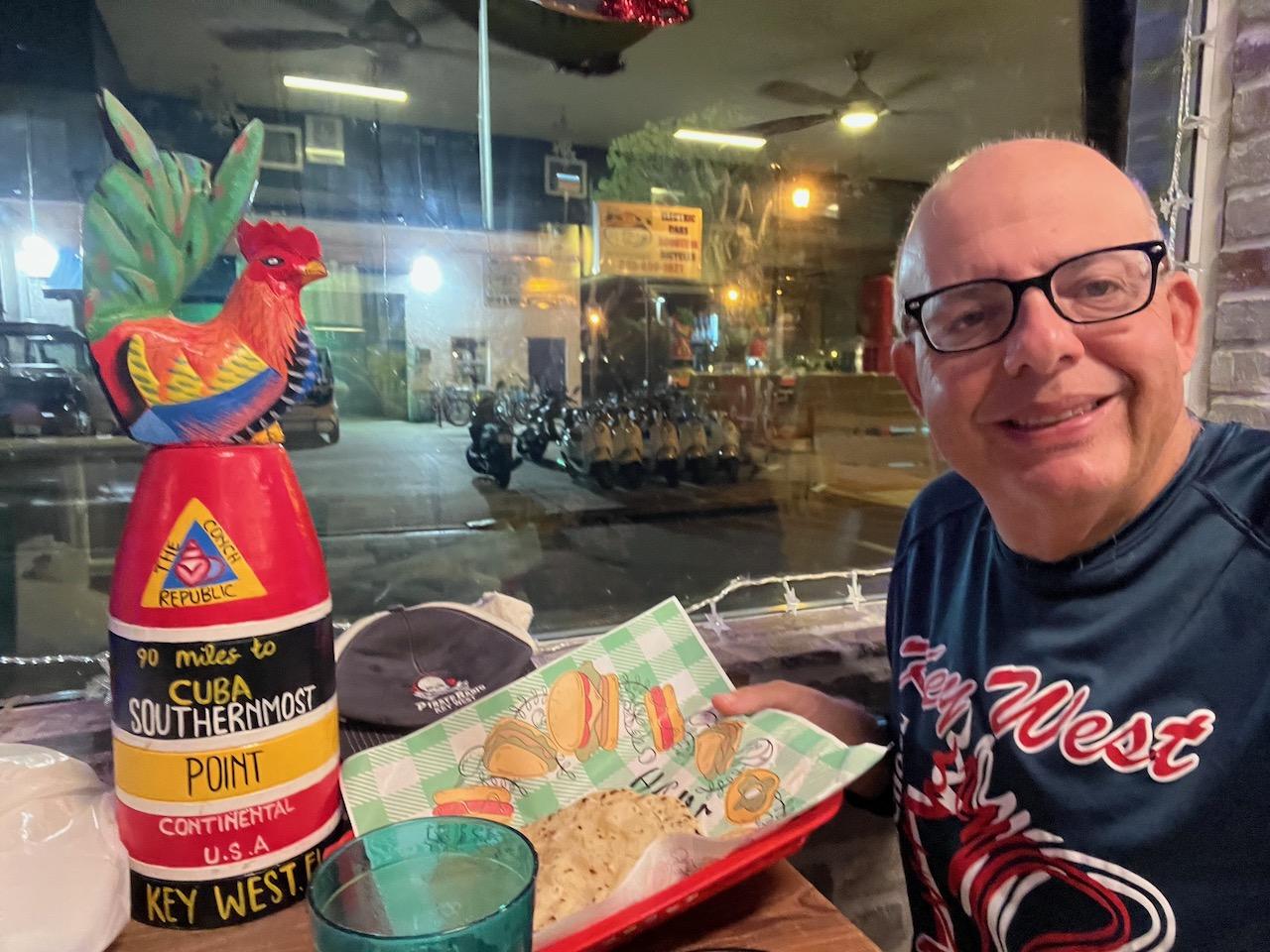 Man smiling at a restaurant table with a colorful rooster sculpture.