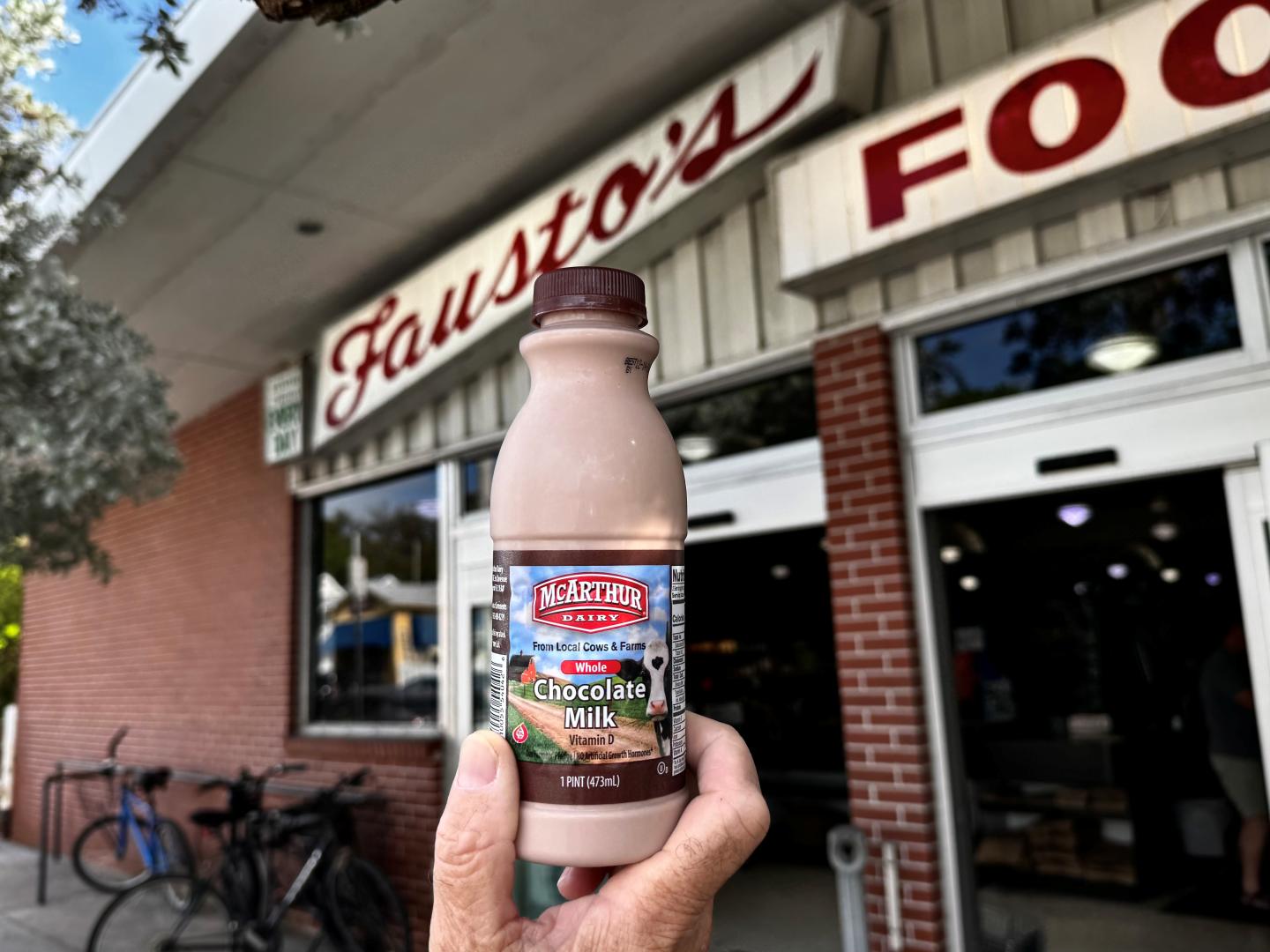 Hand holding chocolate milk bottle in front of Faustos grocery store entrance.