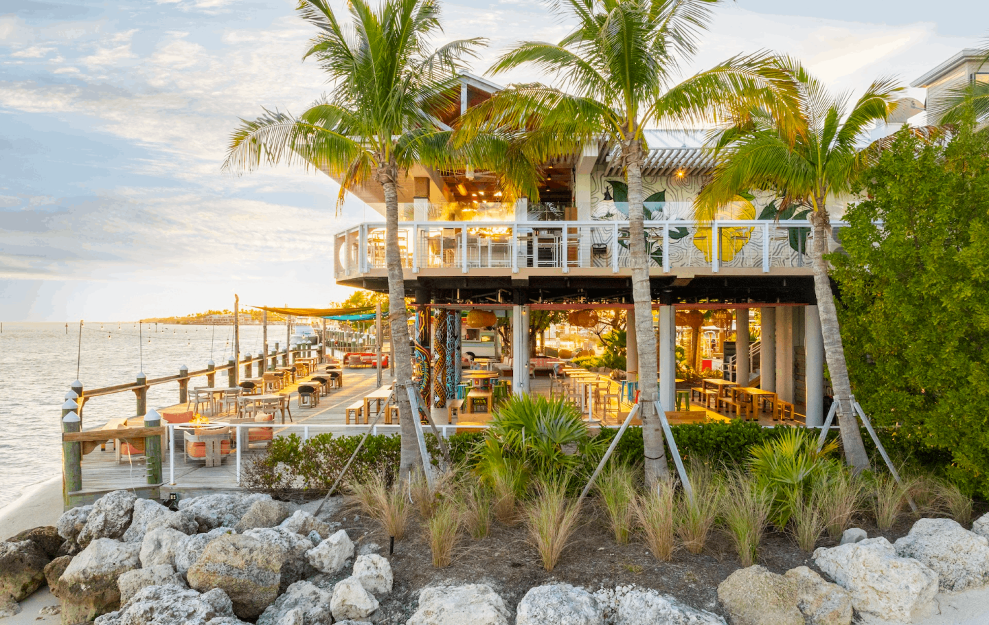 Tropical beachfront restaurant, palm trees, and rocky shore at sunset.