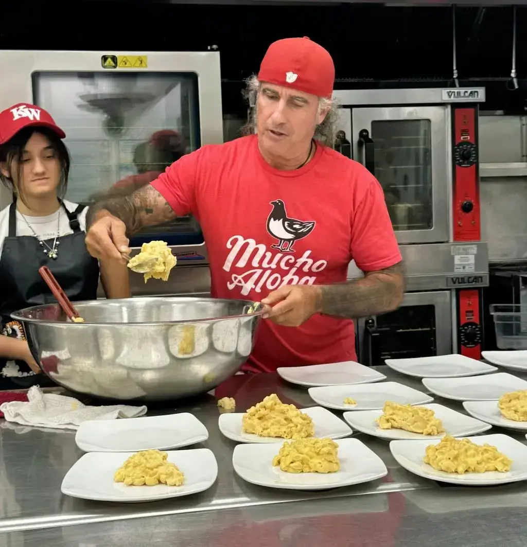 Man in red shirt serving pasta onto plates in a kitchen.