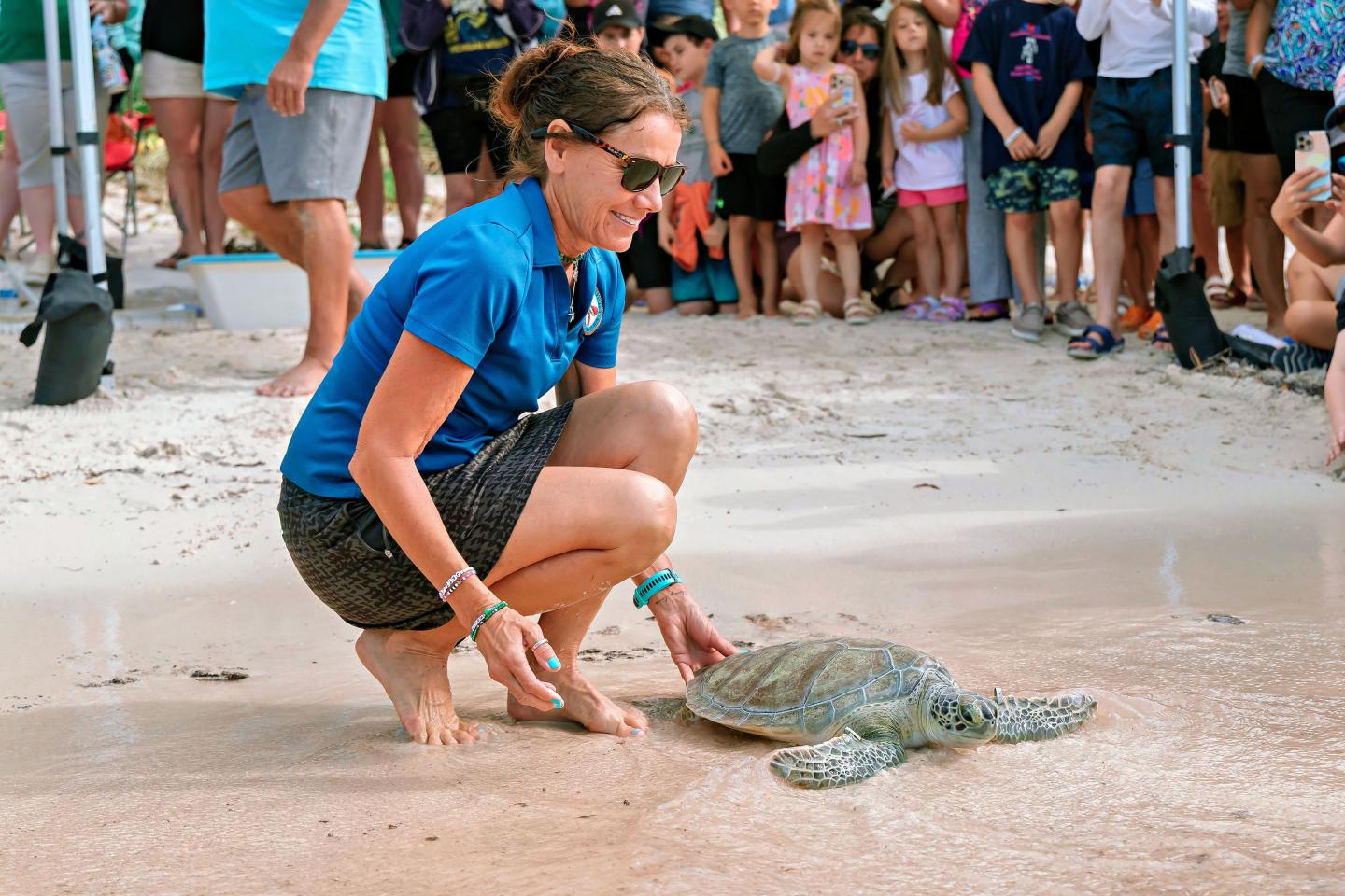 Woman releasing a sea turtle on a beach, crowd watching in the background.