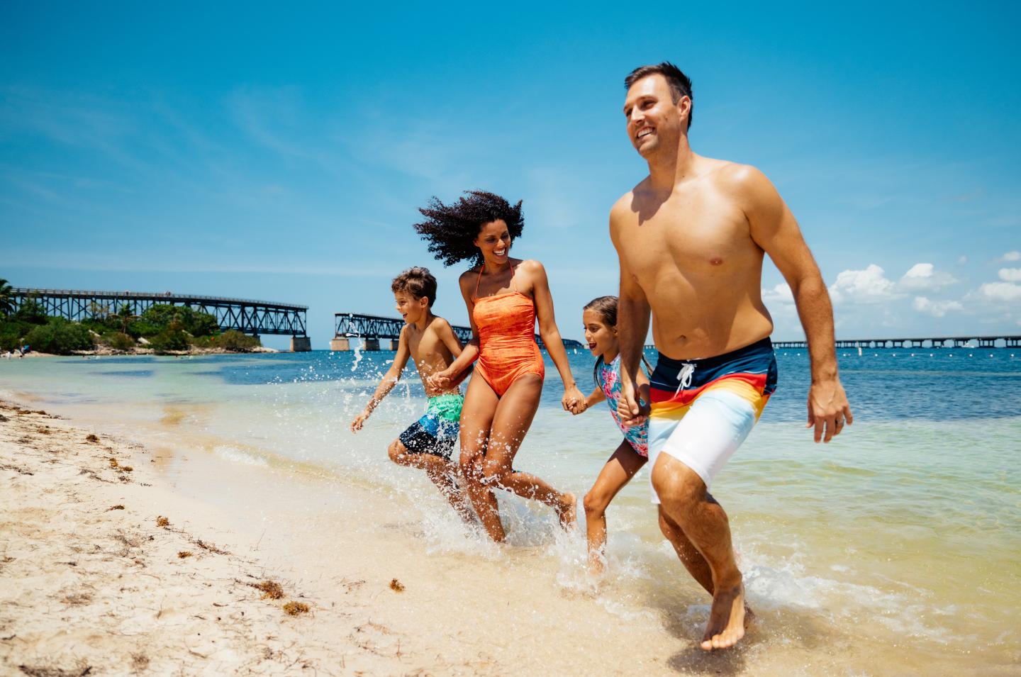 Family running on a sunny beach near the water, looking joyful.
