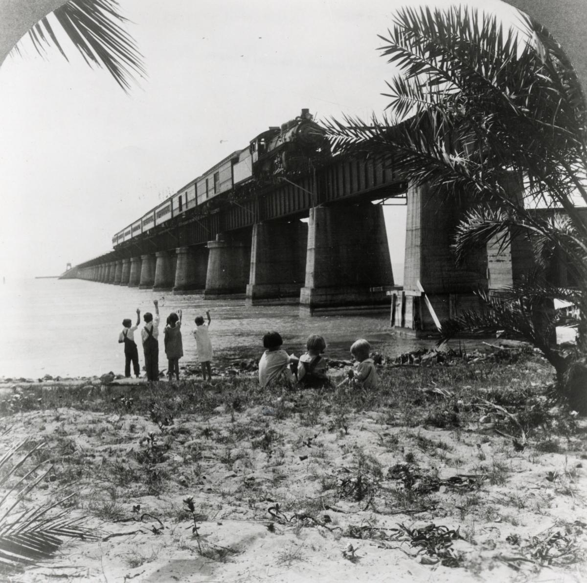 Train crossing a bridge over calm water, with people watching from a sandy shore.
