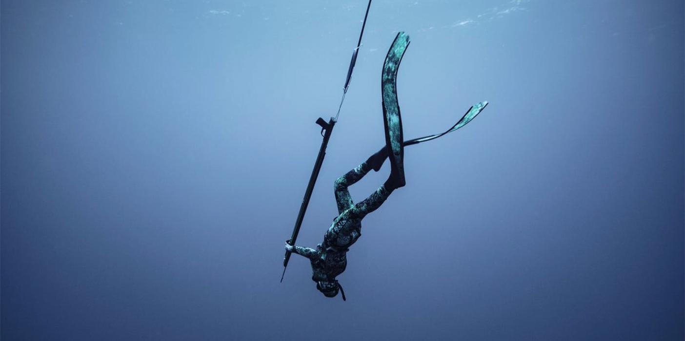 Diver in wetsuit descending underwater, holding a spear.