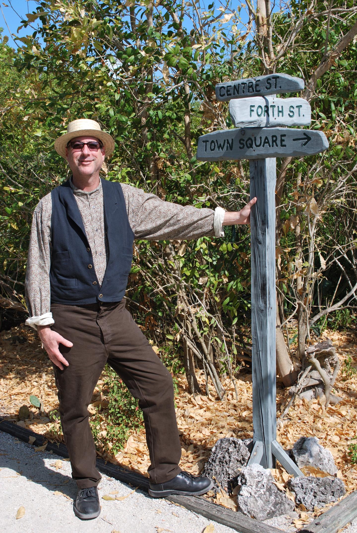 Man in vintage clothing standing by a wooden signpost, outdoors in a sunny setting.