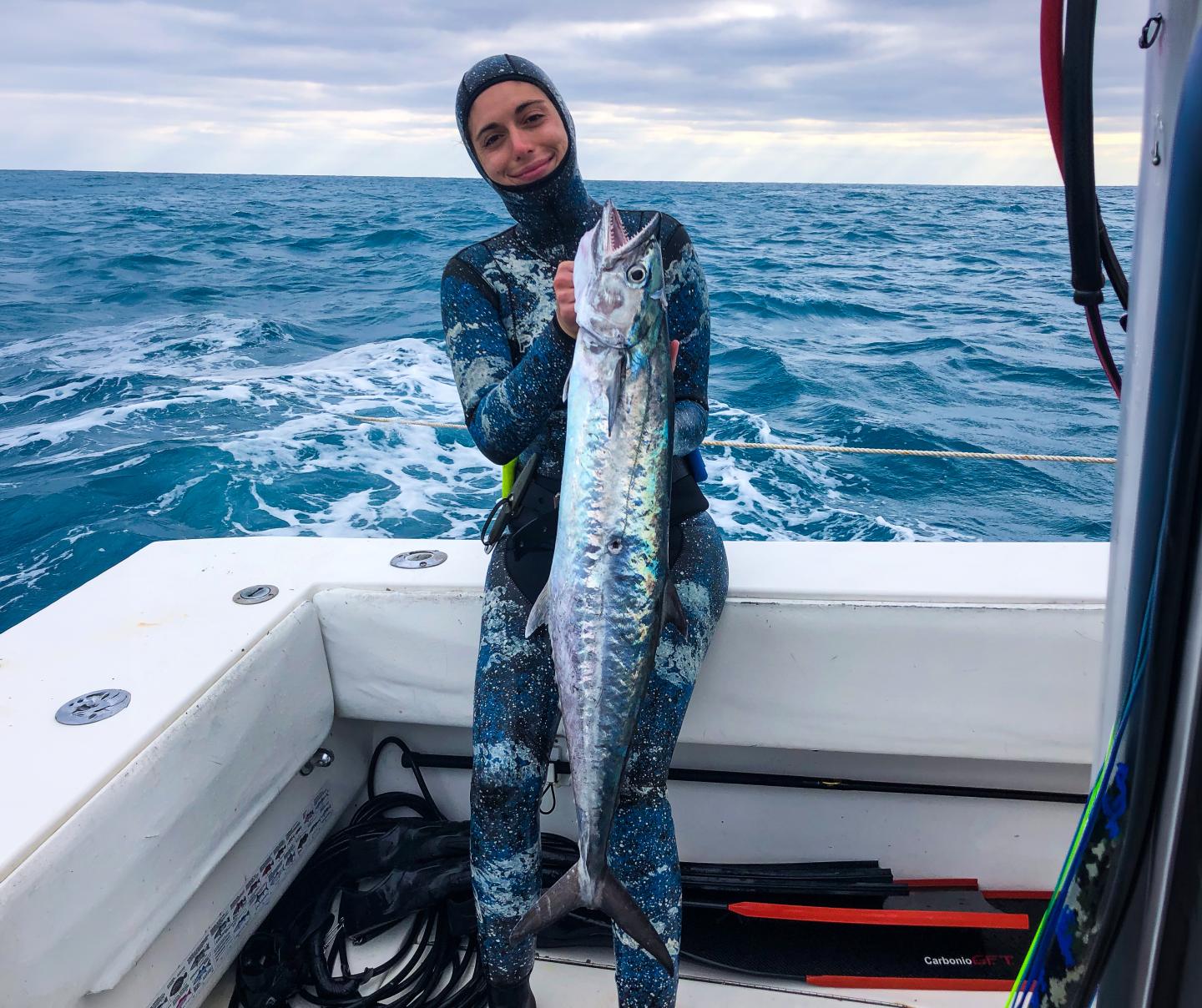 Person in wetsuit holding a large fish on a boat in the ocean.