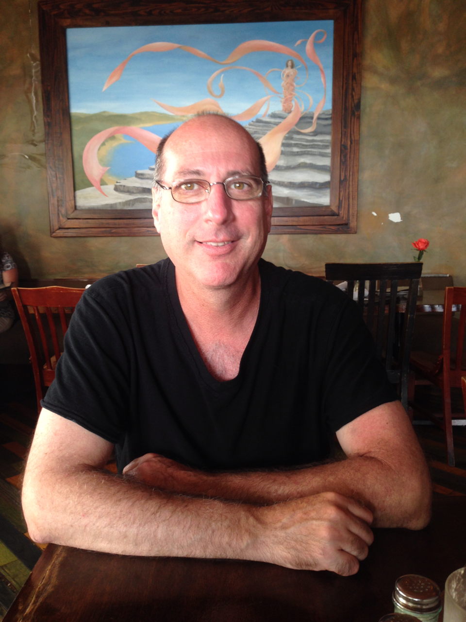 Man in black shirt seated at cafe table, colorful abstract painting behind.