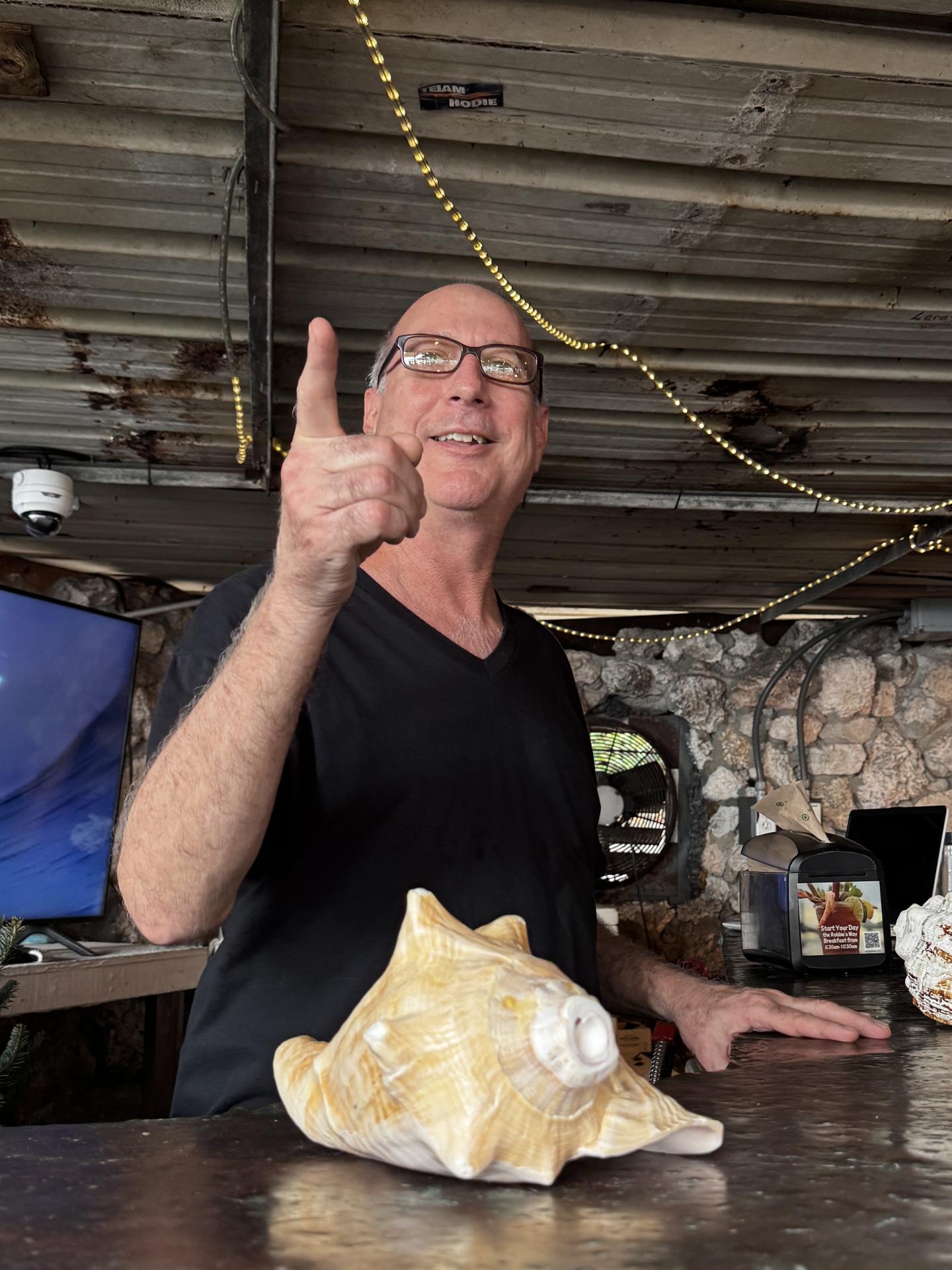 Man smiling, pointing, standing by large conch shells indoors.
