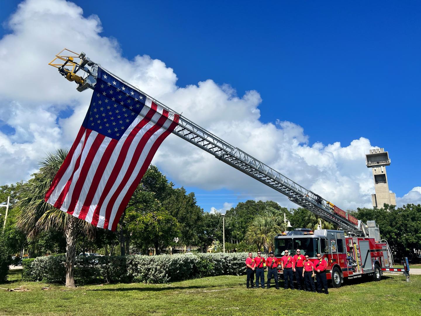 Fire truck with an American flag; firefighters stand nearby under a clear blue sky.