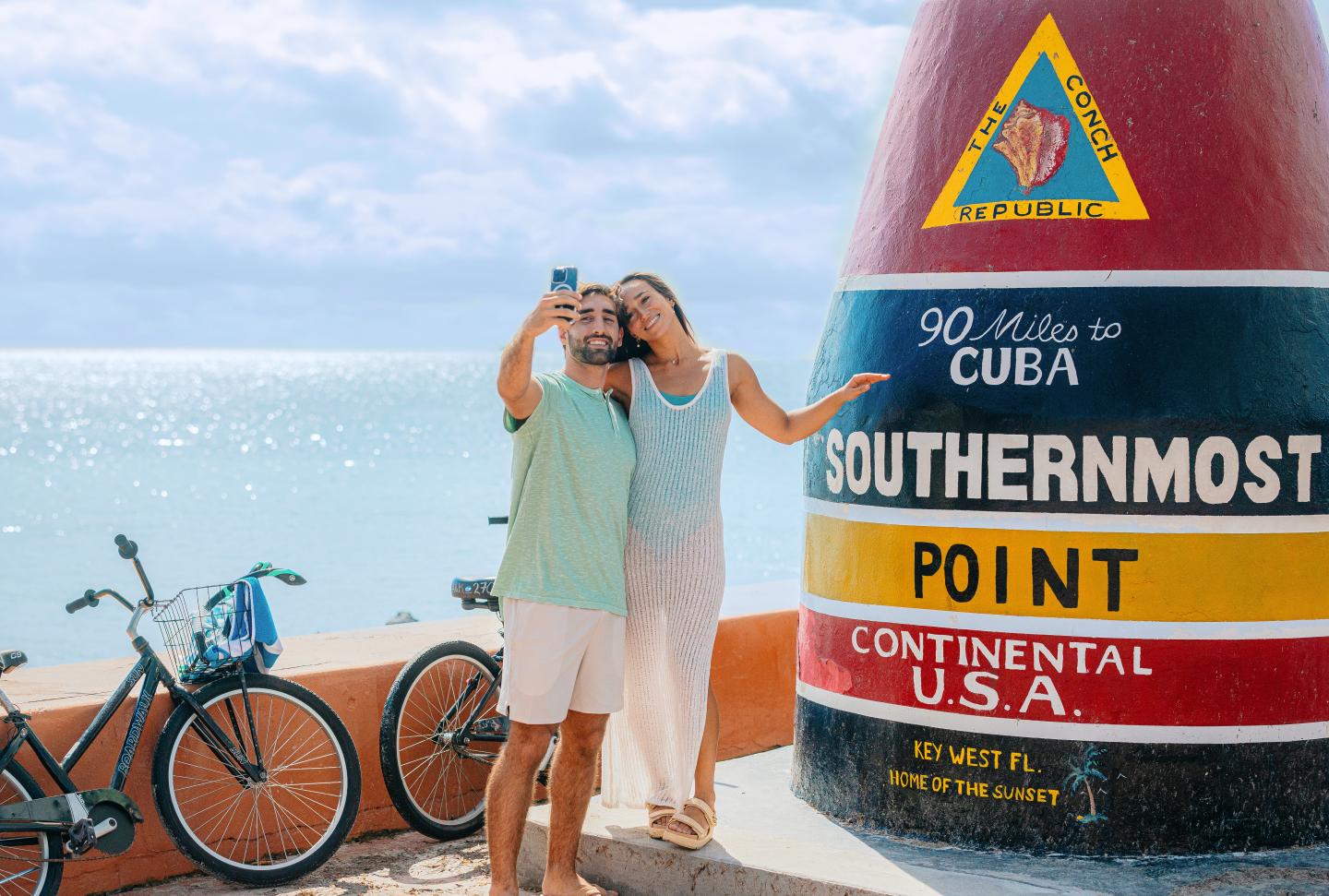 Couple taking a selfie by Southernmost Point buoy, ocean in background.