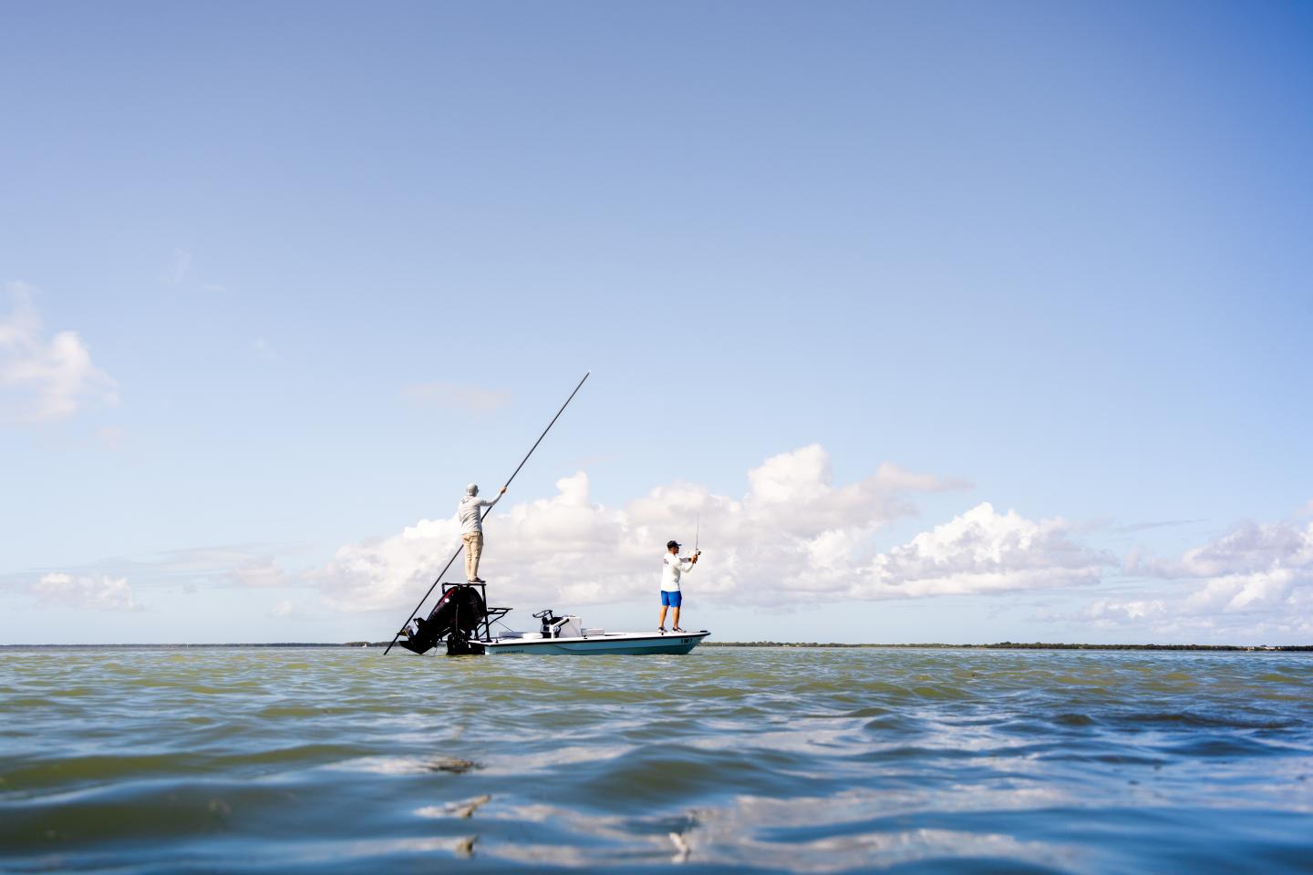 Two people standing on a small boat under a clear blue sky, fishing in calm waters.
