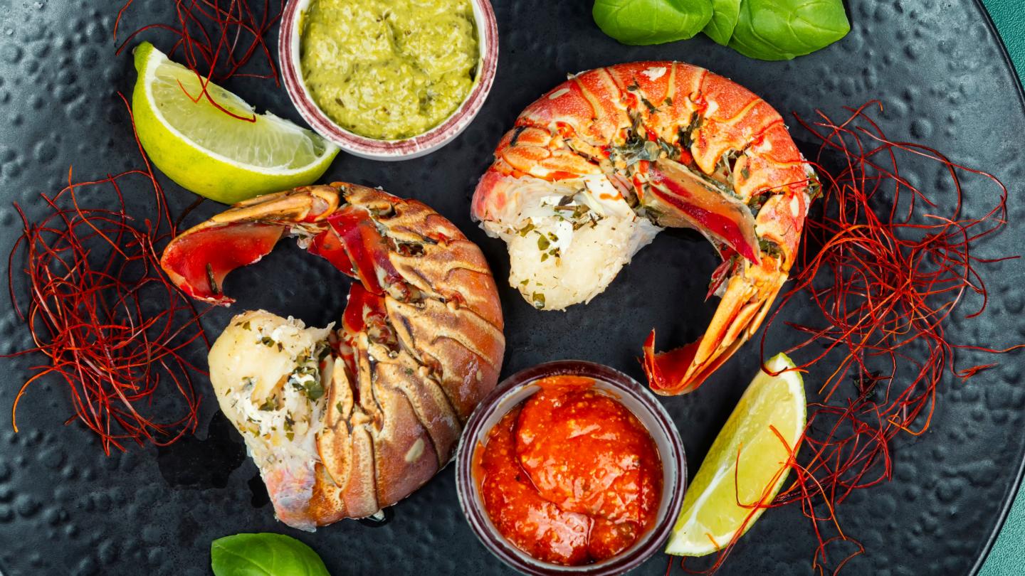 Lobster tails with green and red sauces, lime wedges, and herbs on a dark plate.
