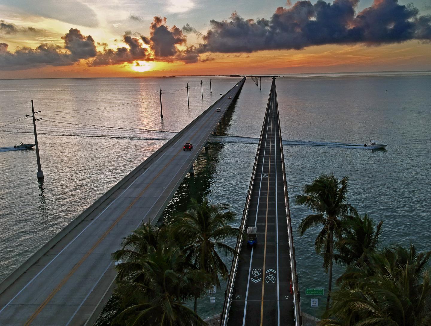 Sunset over a parallel highway bridge with palm trees and boats.