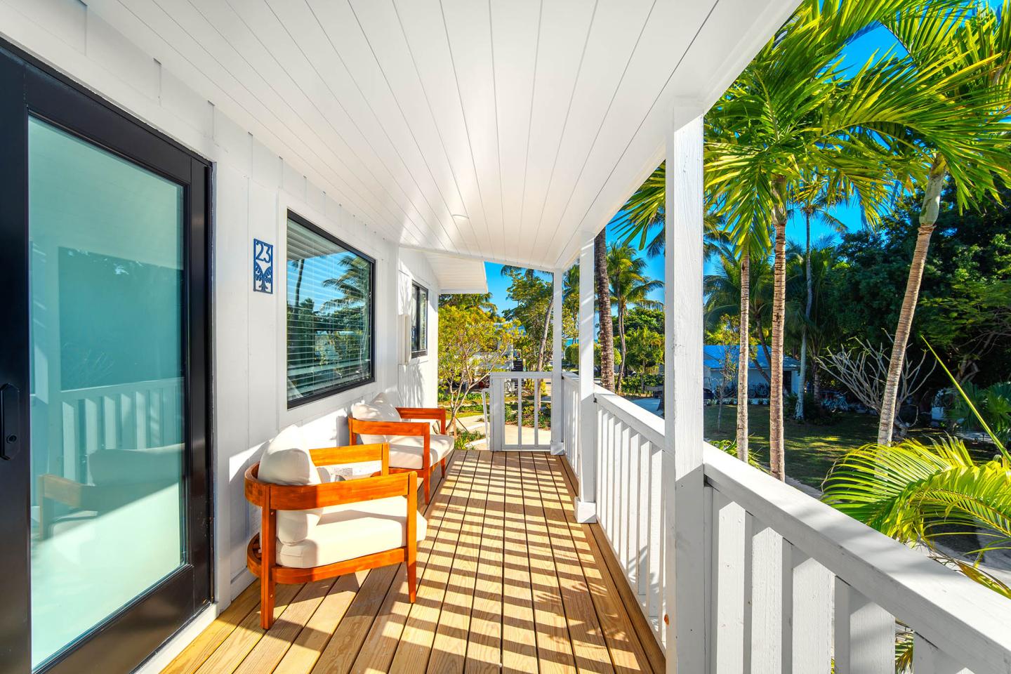 Sunny porch with wooden chairs, white railing, and palm trees nearby.