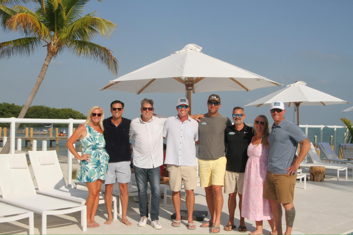 Group of people smiling under beach umbrellas on a sunny day.