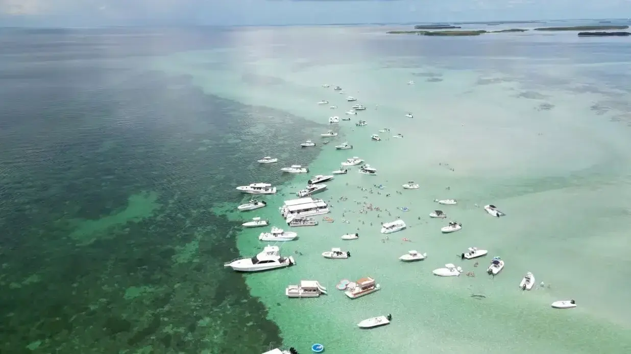 Floating boats on clear turquoise water near a sandbar, aerial view.