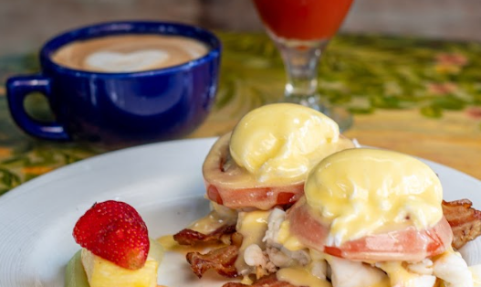 Eggs Benedict with hollandaise sauce, cappuccino, and a bloody mary on a colorful table.
