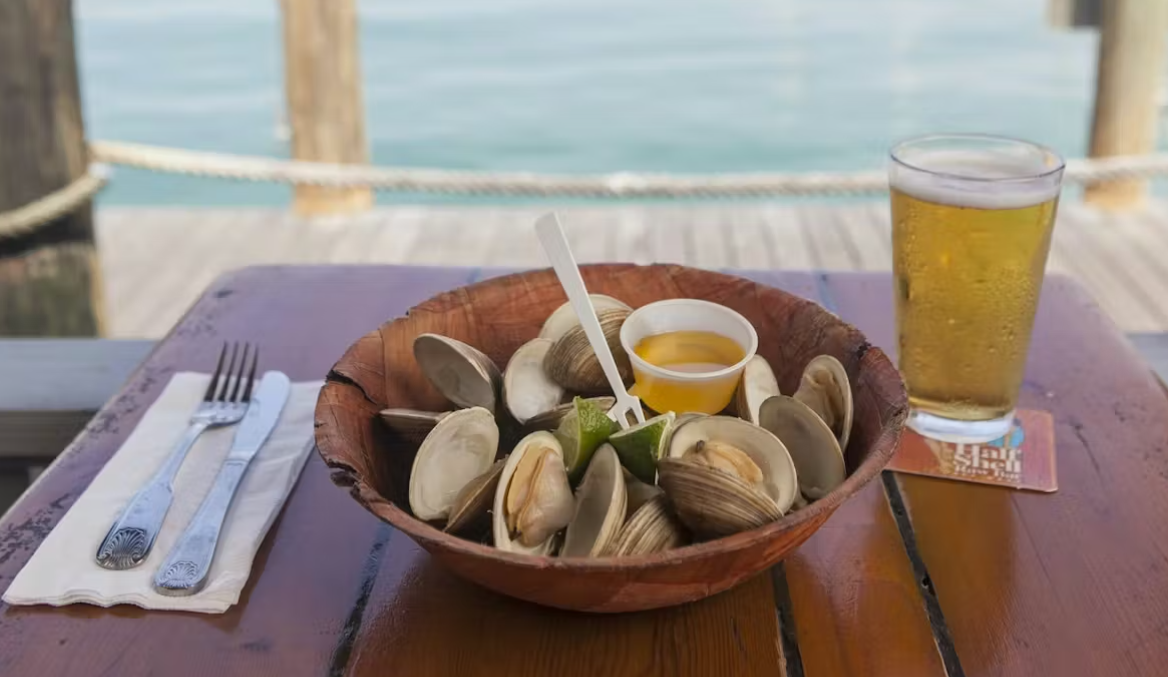 Clams and dip with lime in a wooden bowl, a beer on a dockside table.