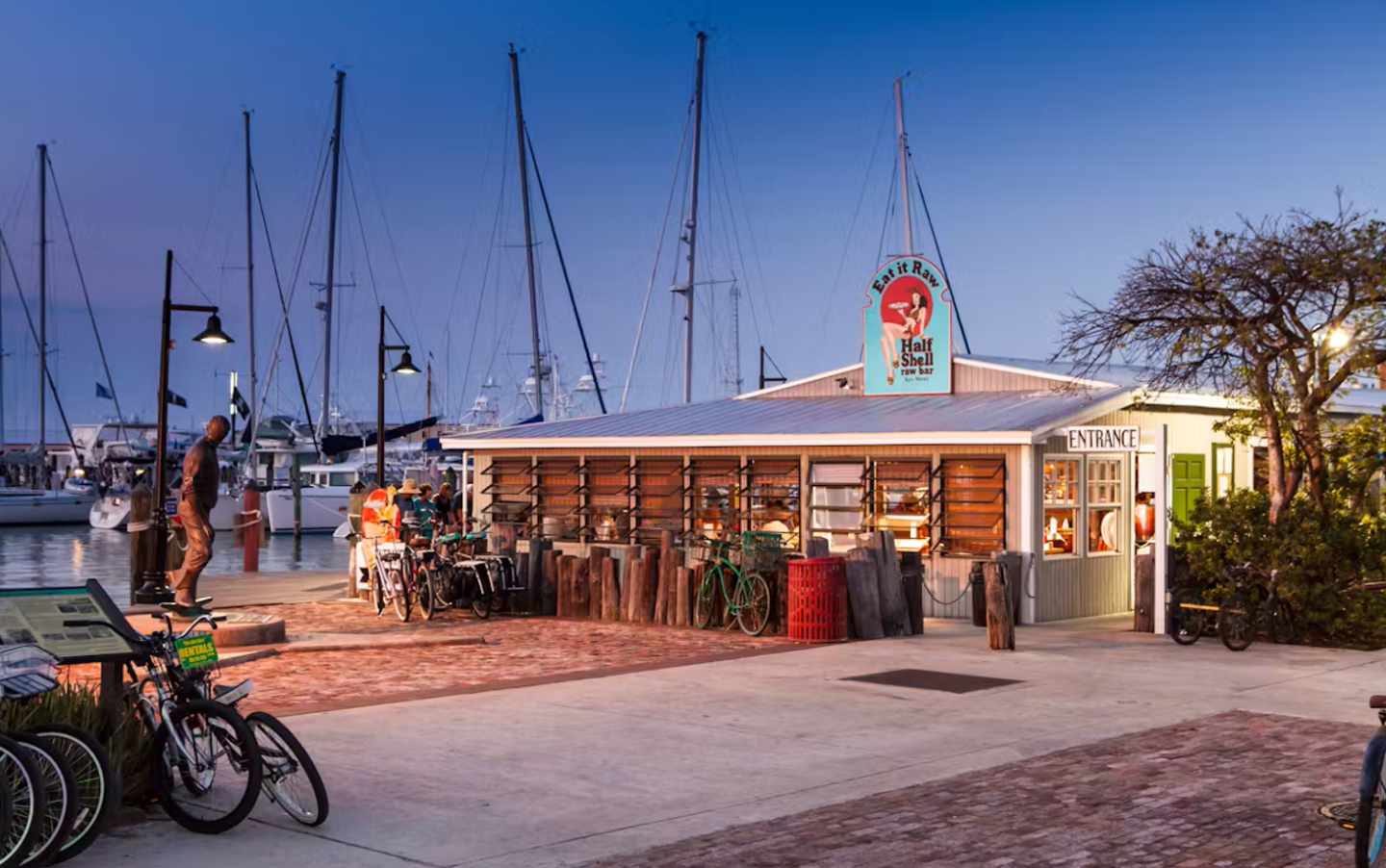 Cozy waterfront café at sunset, bicycles parked outside, sailboats in the background.