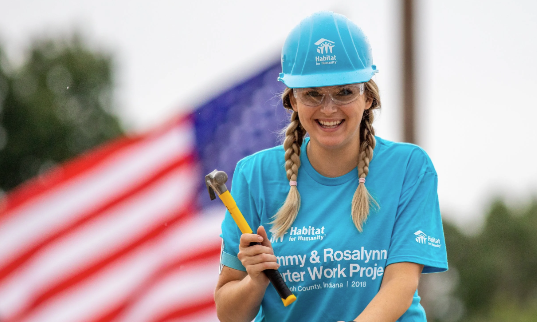 Smiling woman in a blue hard hat holding a hammer, with an American flag in the background.